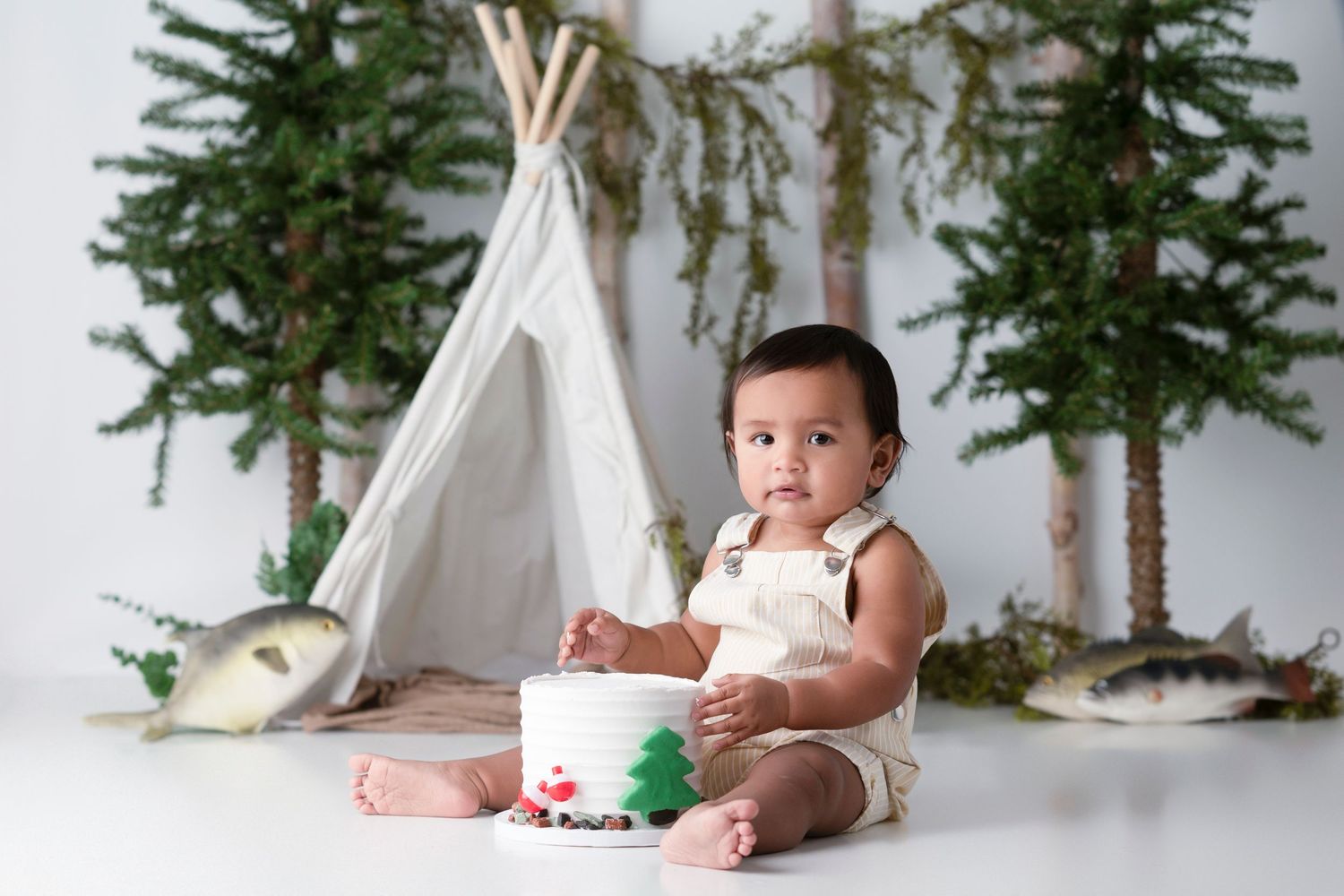 Baby in white onesie lies on white blanket with evergreen trees and teepee decorations in background.