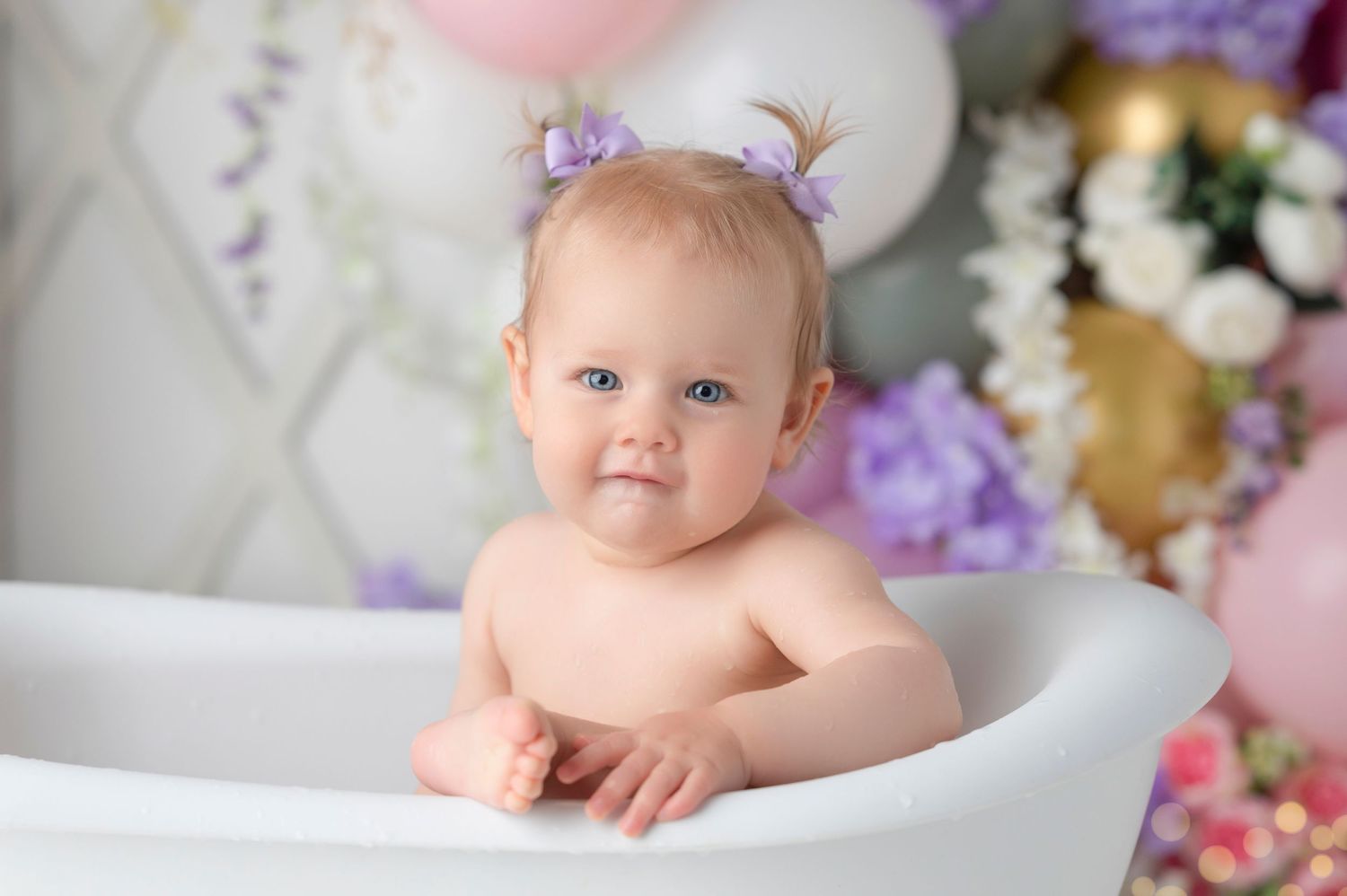 A cute baby girl with purple bows sits in a white mini bathtub surrounded by pastel balloons and flowers.
