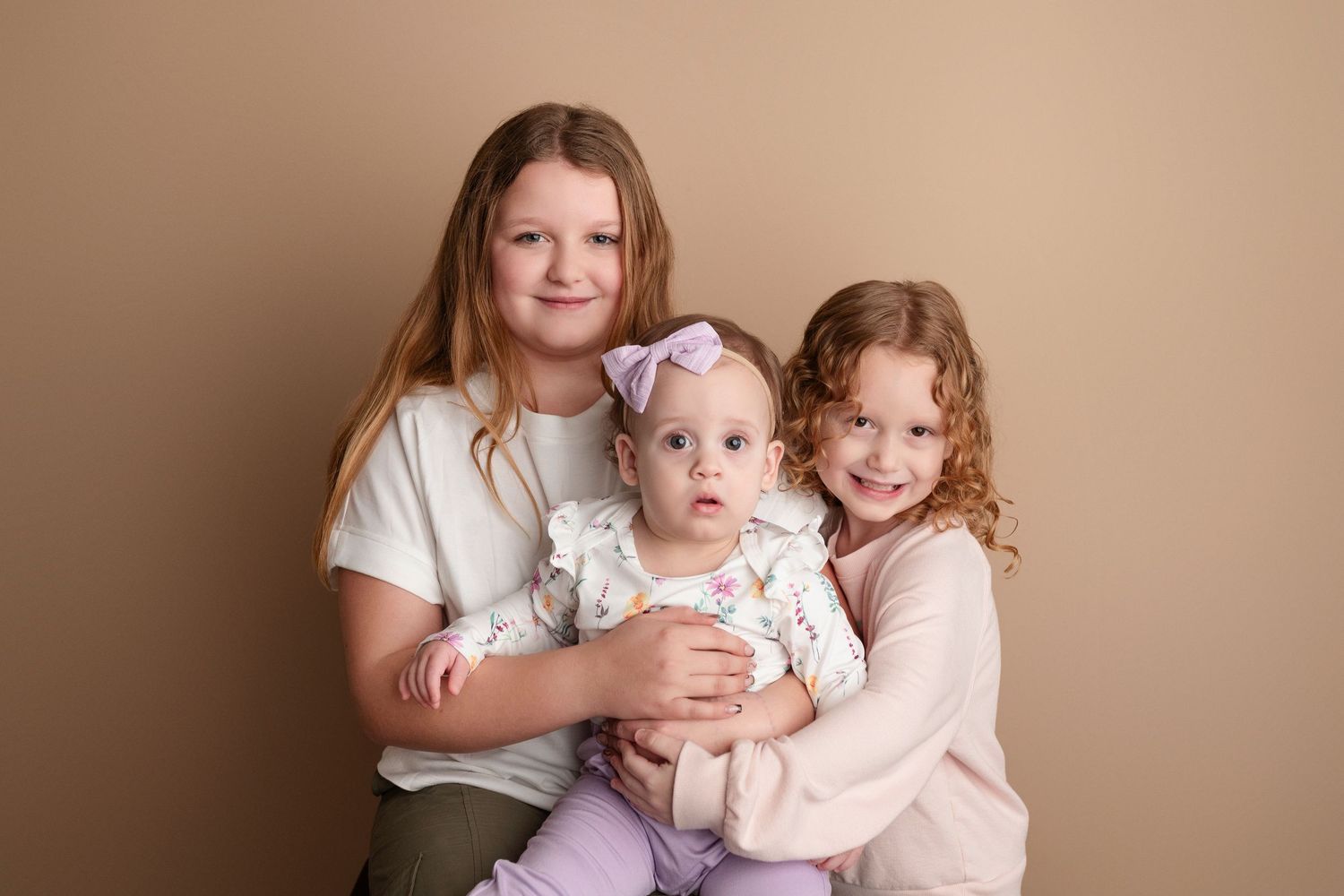 Two young girls pose with a baby in a purple headband for a studio portrait on a beige background.