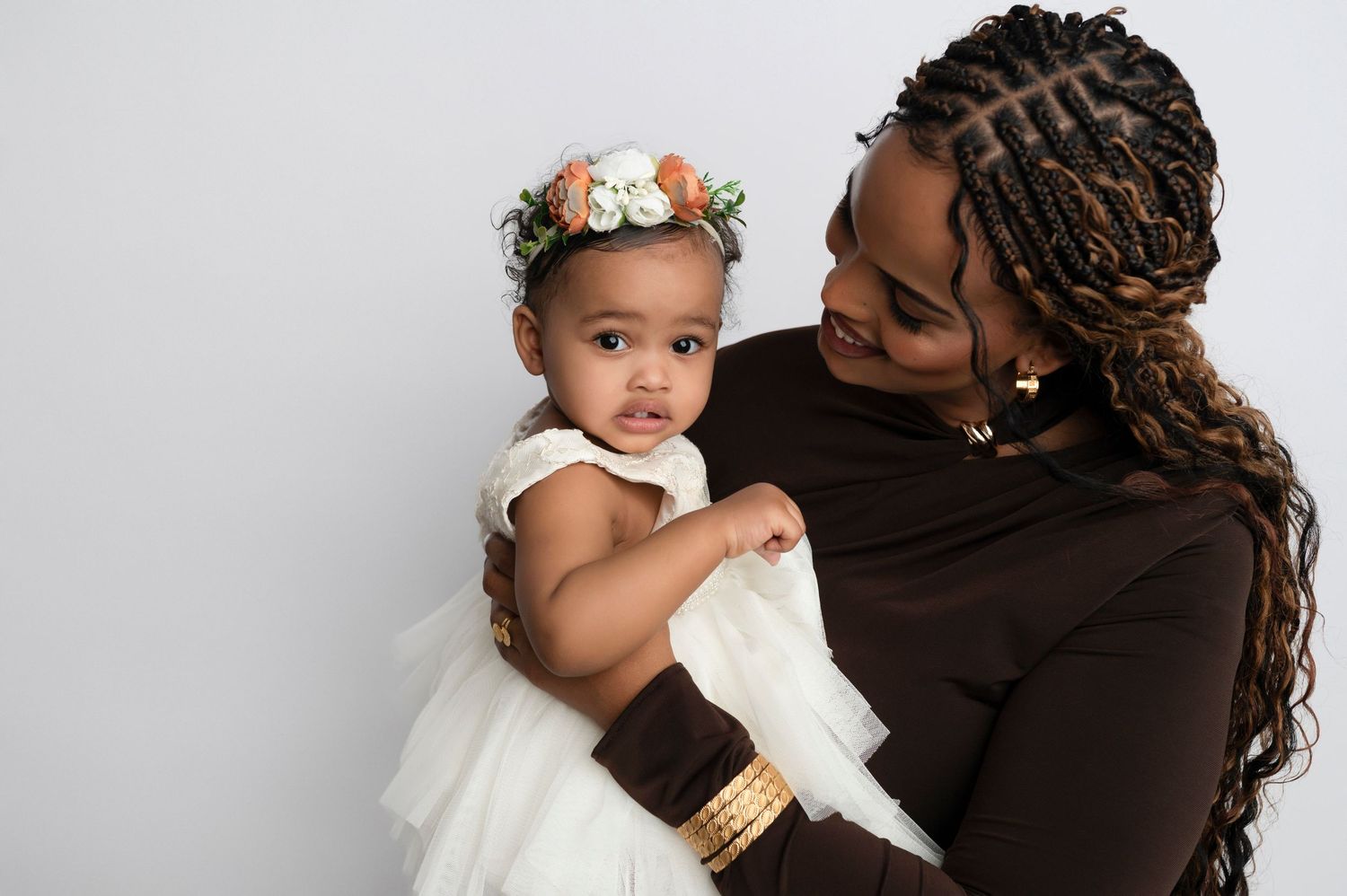 A mother in brown holds her baby girl in a white dress and floral crown against a light gray background.