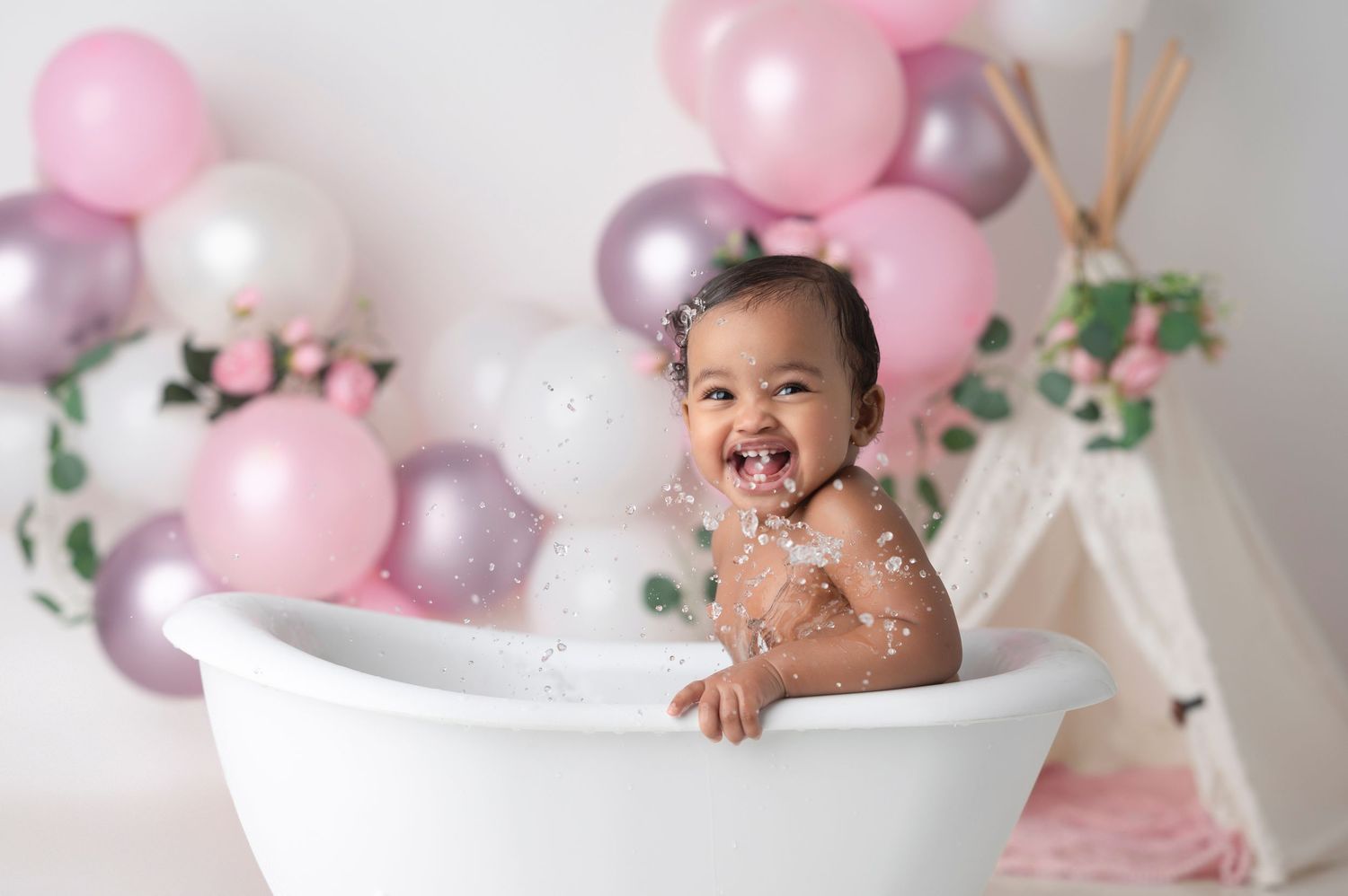Happy baby girl splashing in a white bathtub surrounded by pink and silver balloons during a first birthday cake smash photo session.