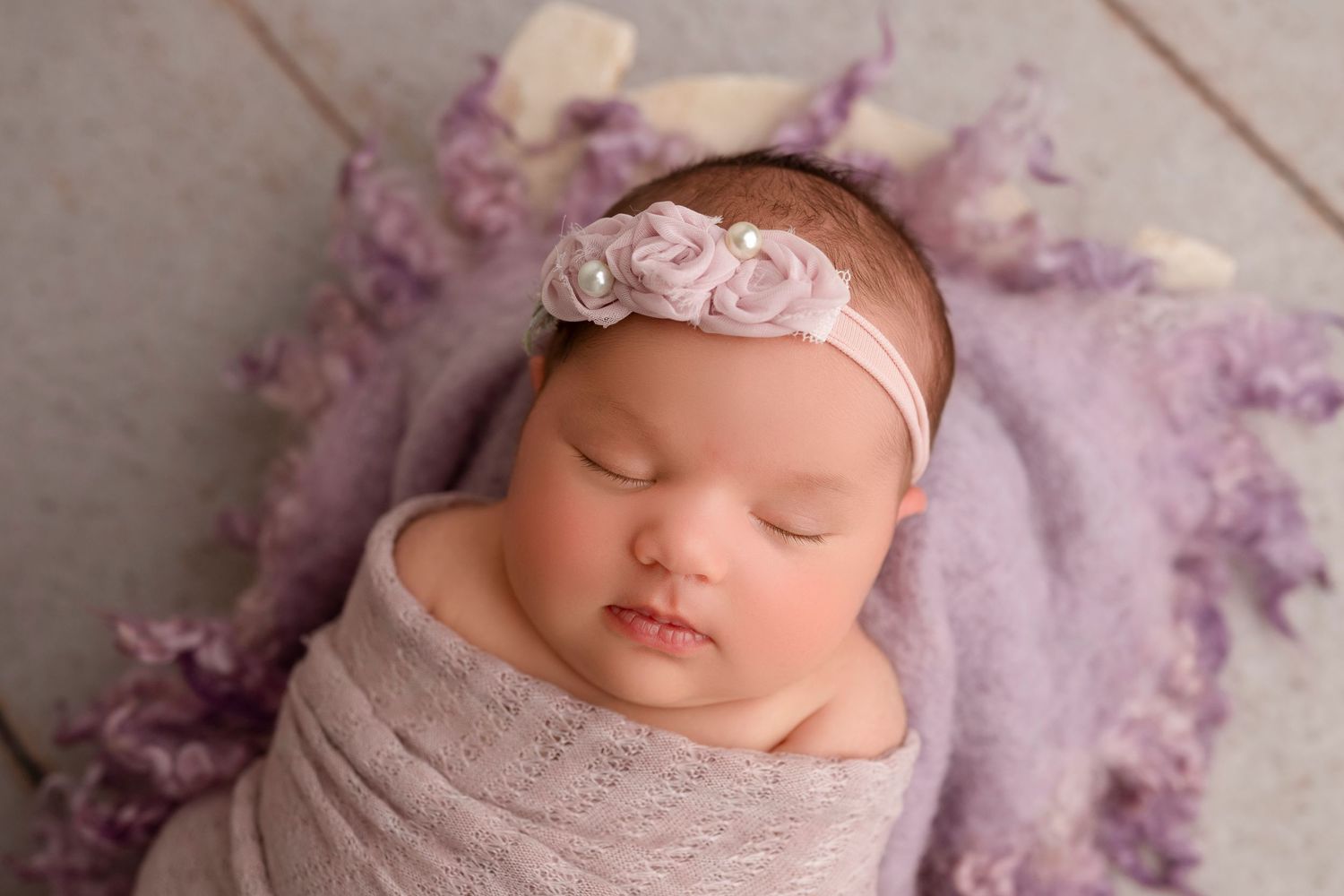 Sleeping newborn baby wrapped in pink blanket with delicate lace headband lying on soft purple backdrop.