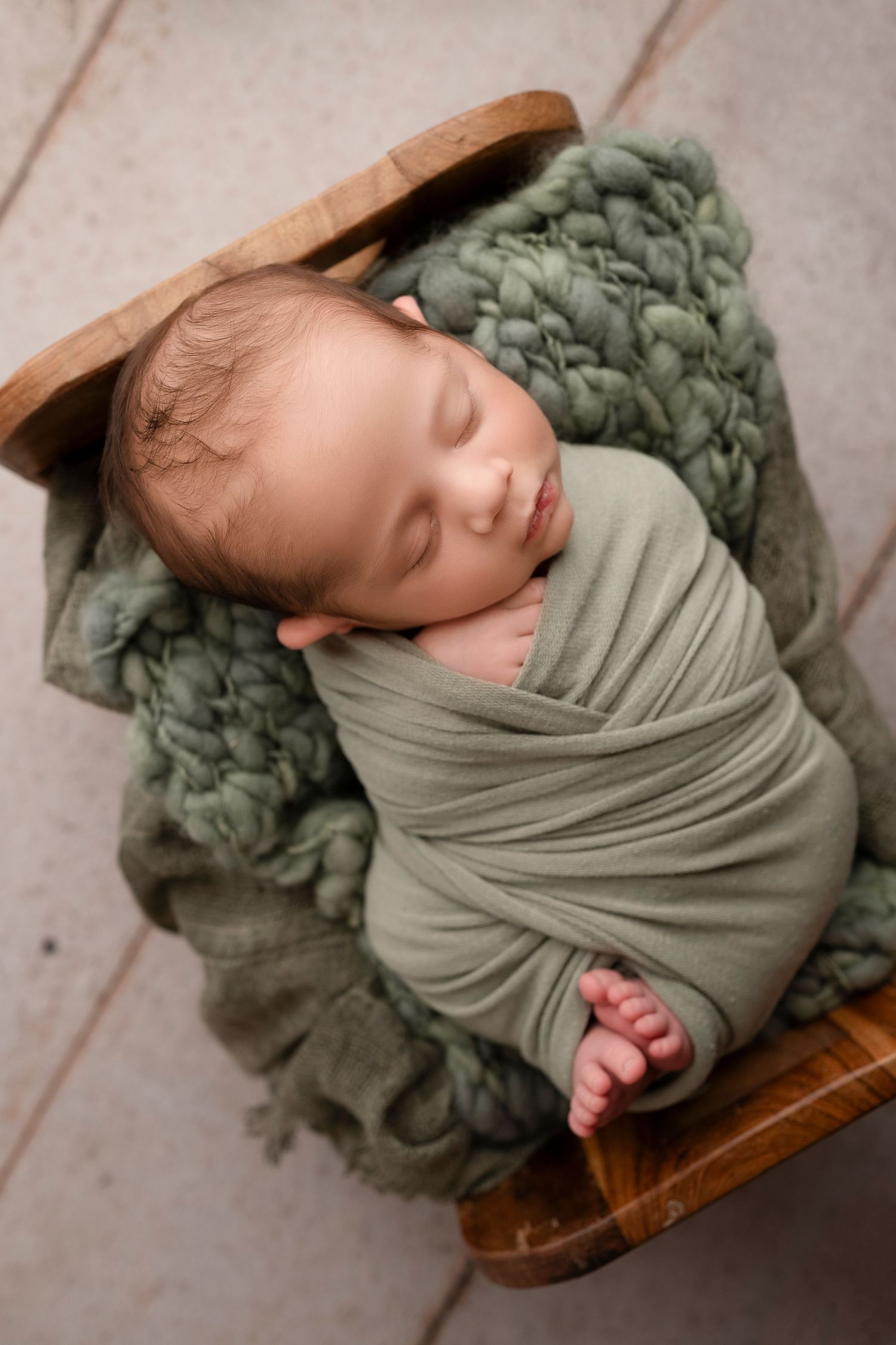 Newborn baby swaddled in olive green fabric resting peacefully on wooden surface in studio setting.