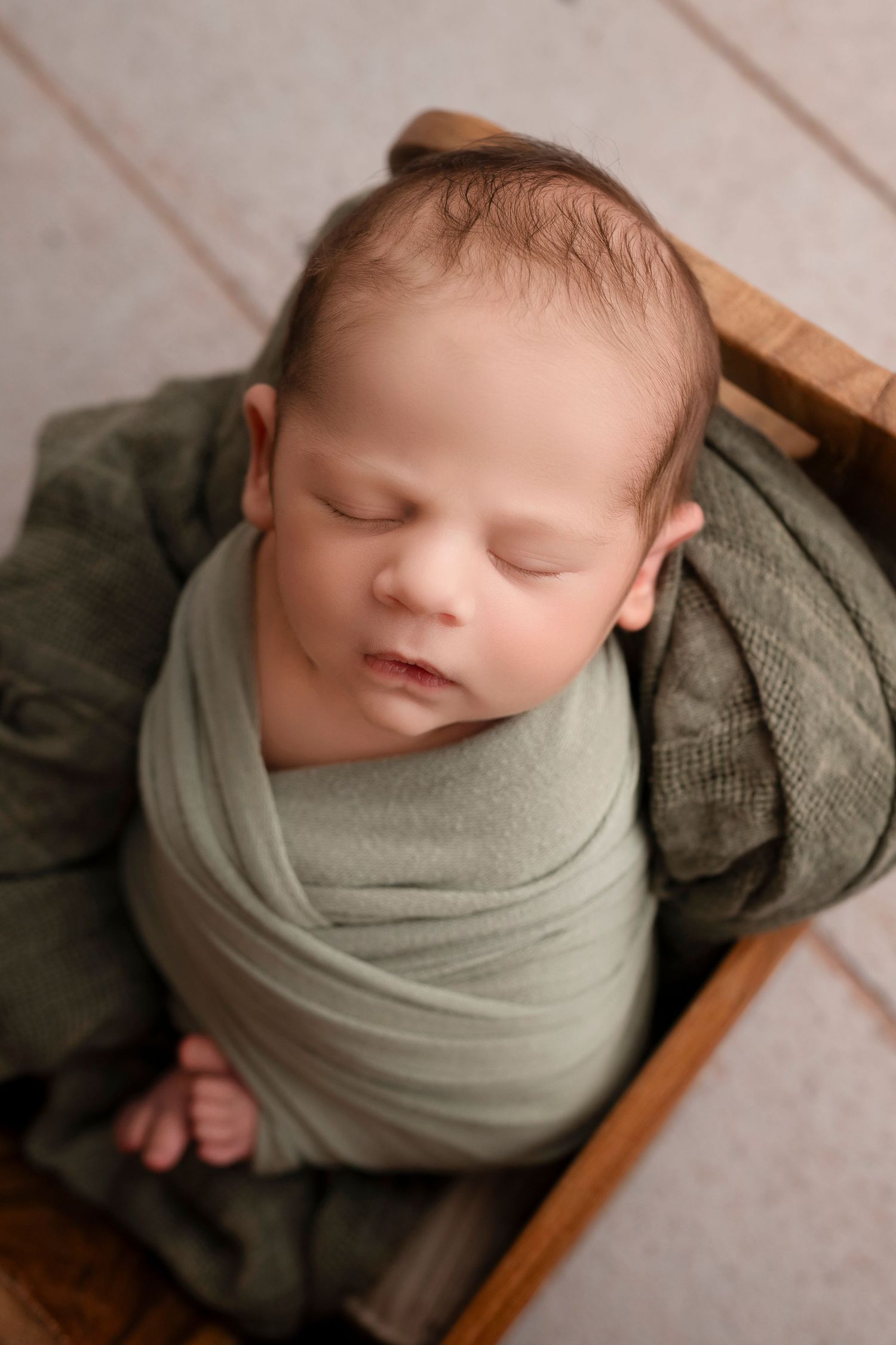 Newborn photography series showing a sleeping baby wrapped in sage green fabric against a light wooden backdrop.