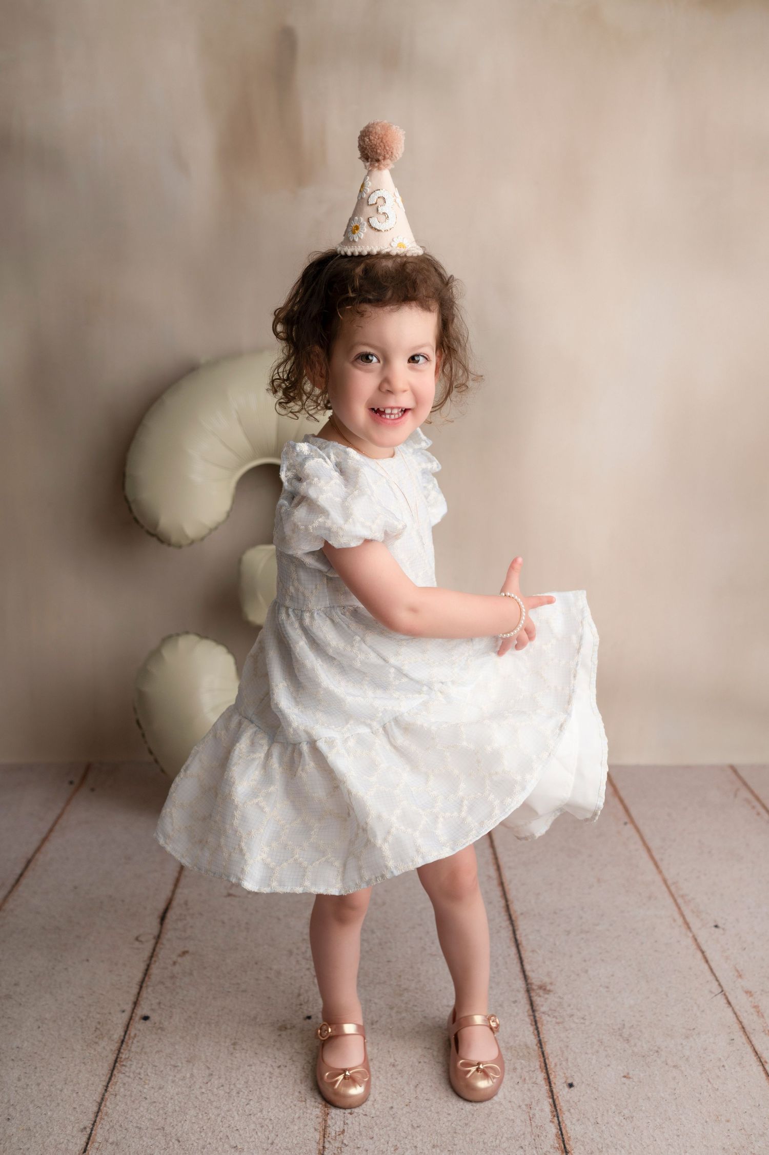 A small child in a white lace dress and brown sandals twirls and plays in a studio setting with cream-colored backdrop.