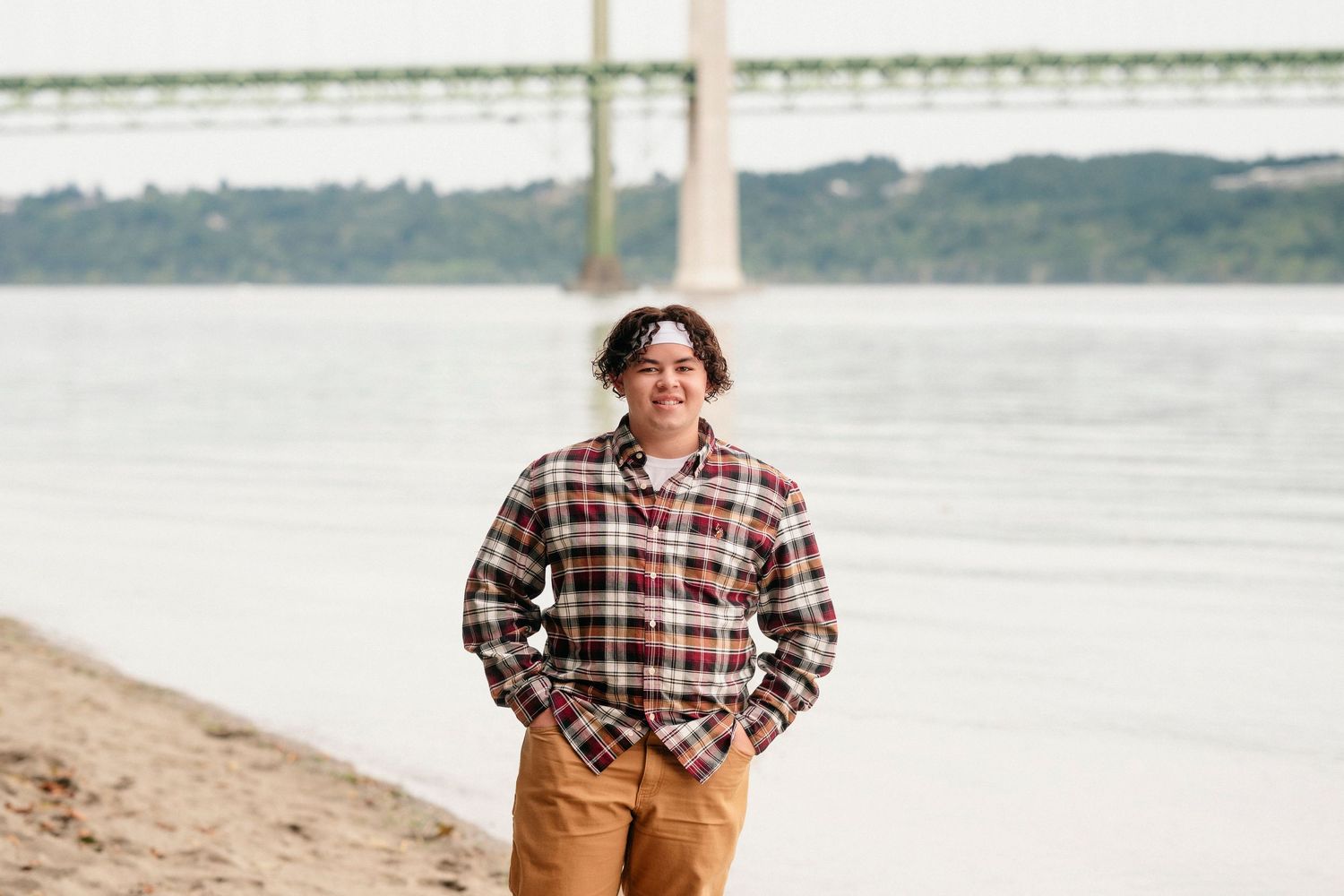 A person in a plaid shirt and brown pants stands on a sandy beach with water and coastline in the background.