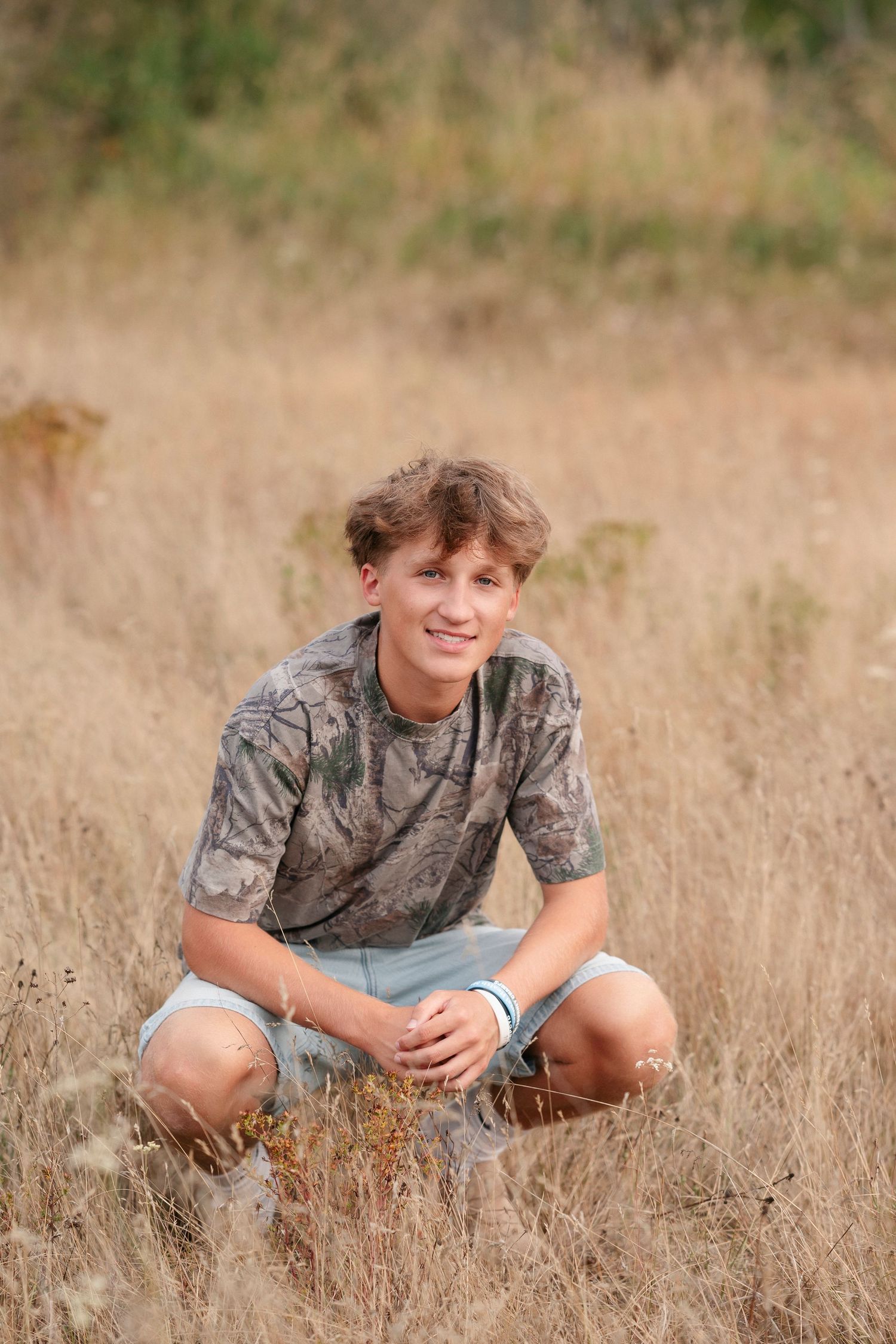 A young person sits in a field of tall dried grass wearing a camouflage t-shirt and denim shorts.