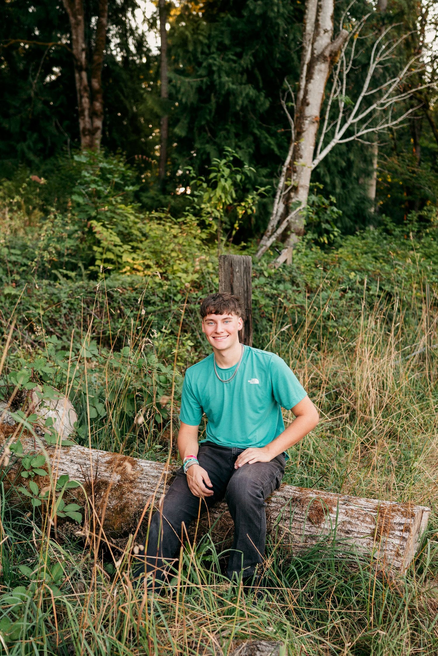 Someone sits casually on a fallen log in a wooded area wearing a teal shirt and dark pants.