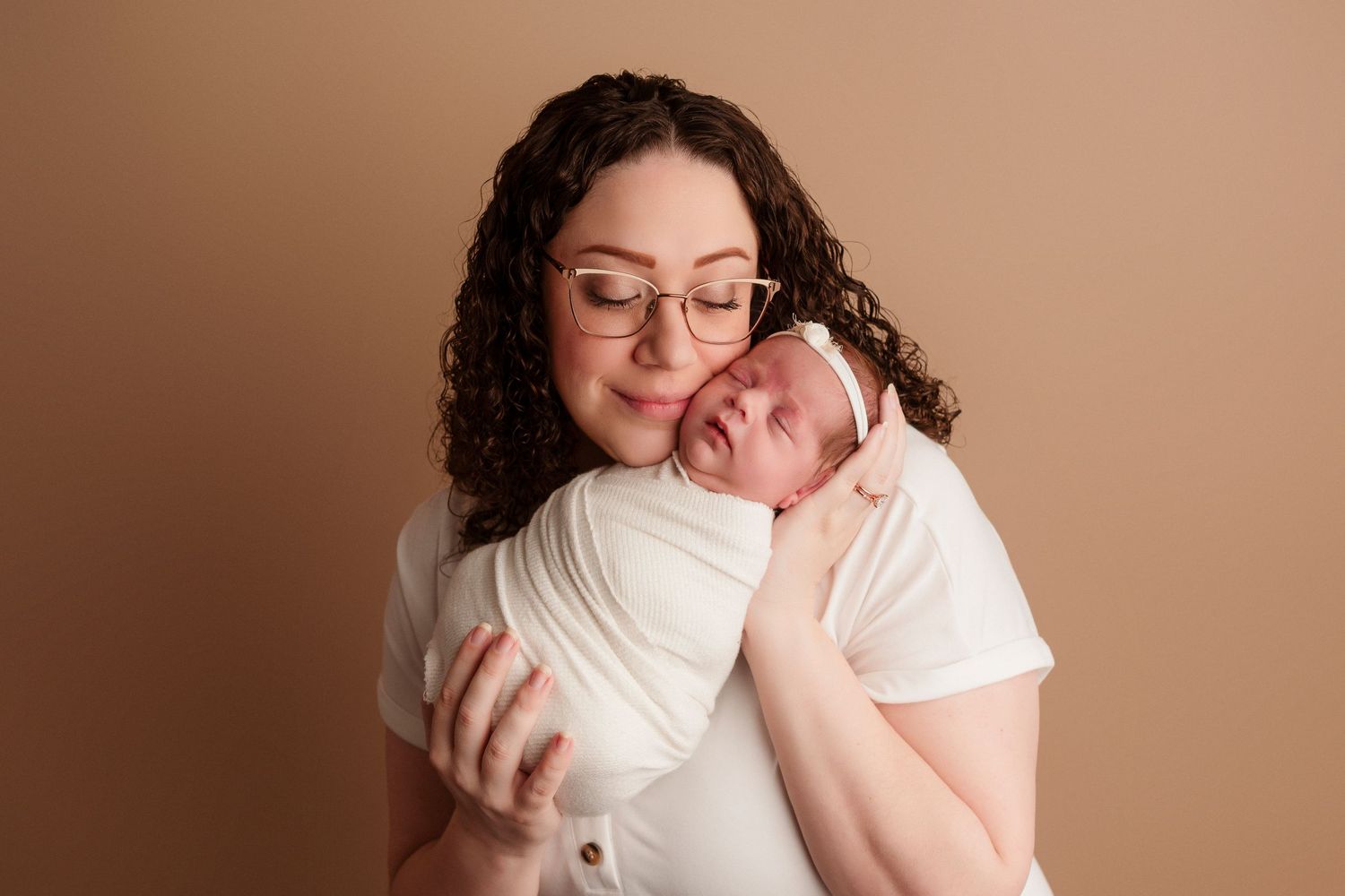 A tender newborn photography session shows a gentle embrace against a warm brown backdrop.