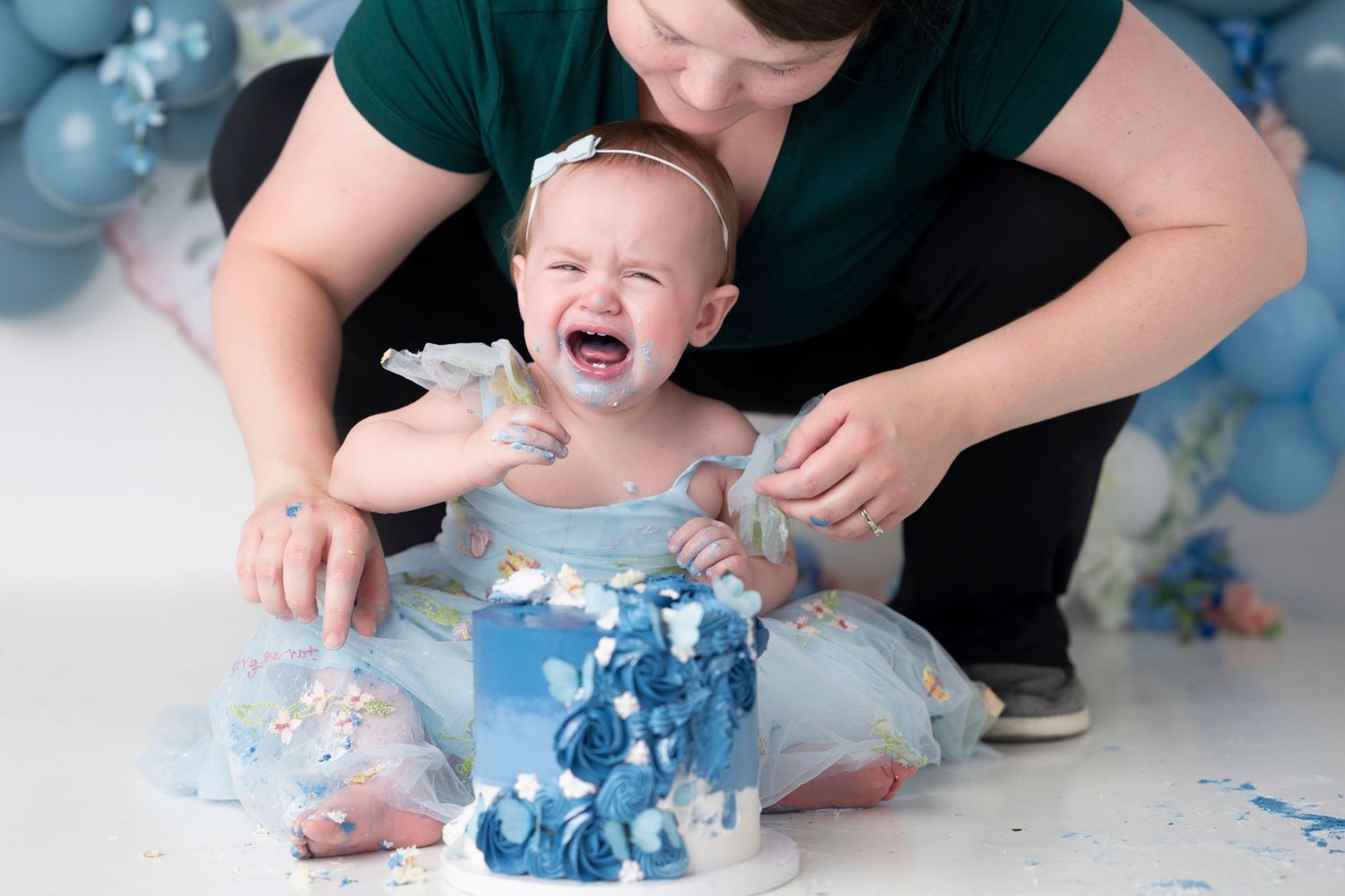 Baby smashing blue cake during first birthday cake smash photoshoot with decorative backdrop.