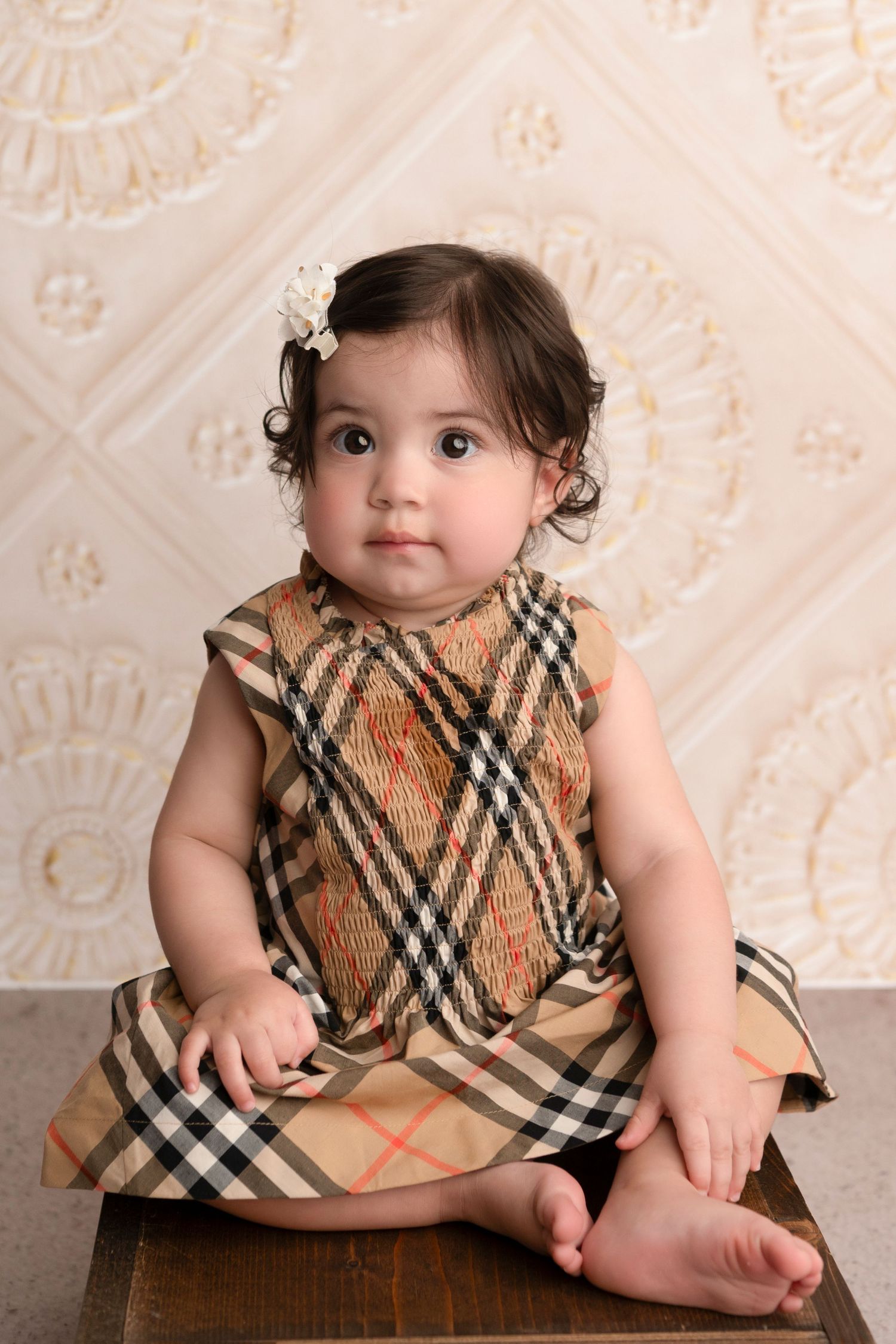 A baby wearing a plaid dress with a floral hair clip sits on a wooden surface against a patterned backdrop.