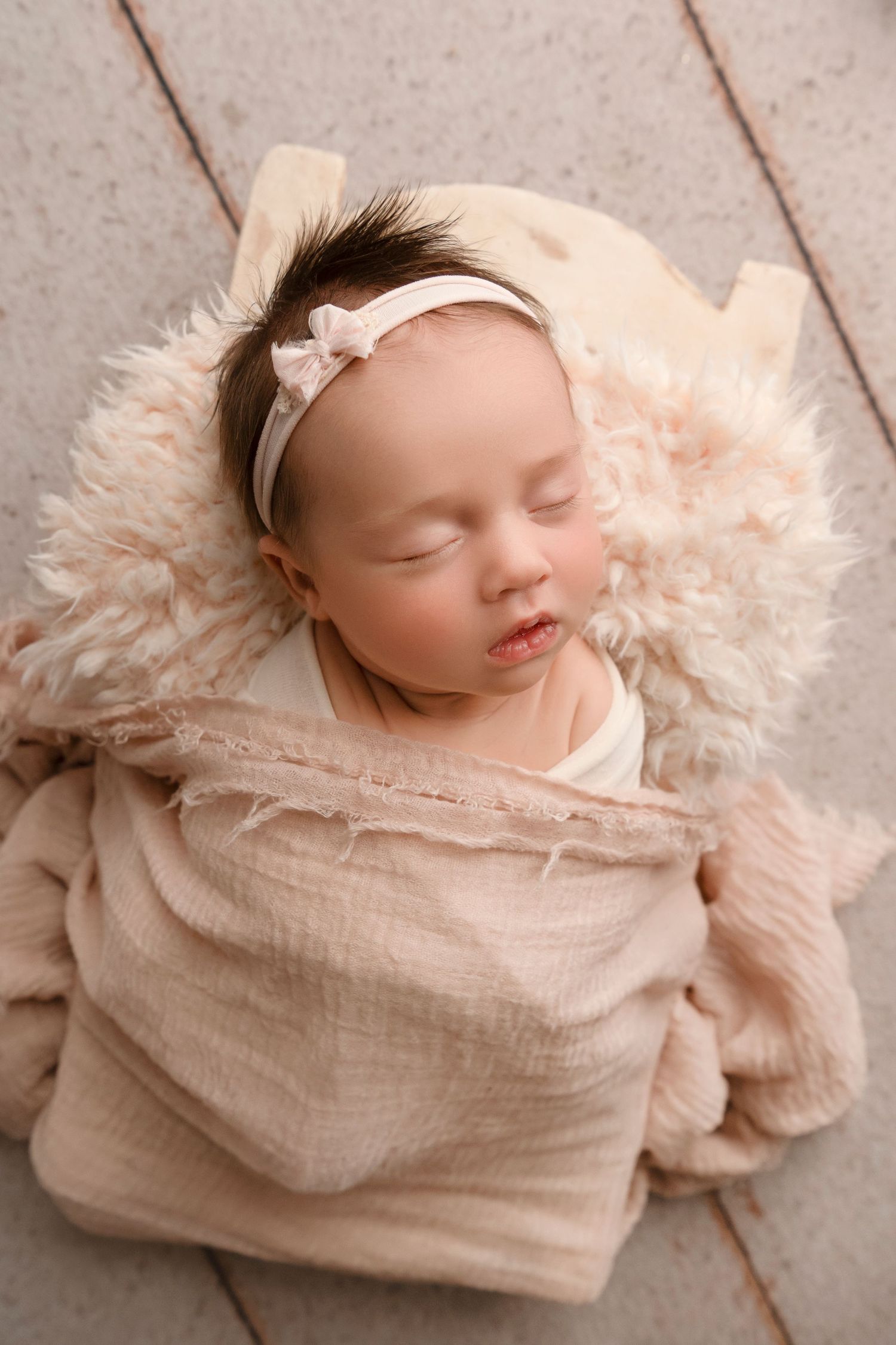 Newborn baby wrapped in soft peach fabric lies peacefully on fluffy cream-colored fur blanket in professional photo series.