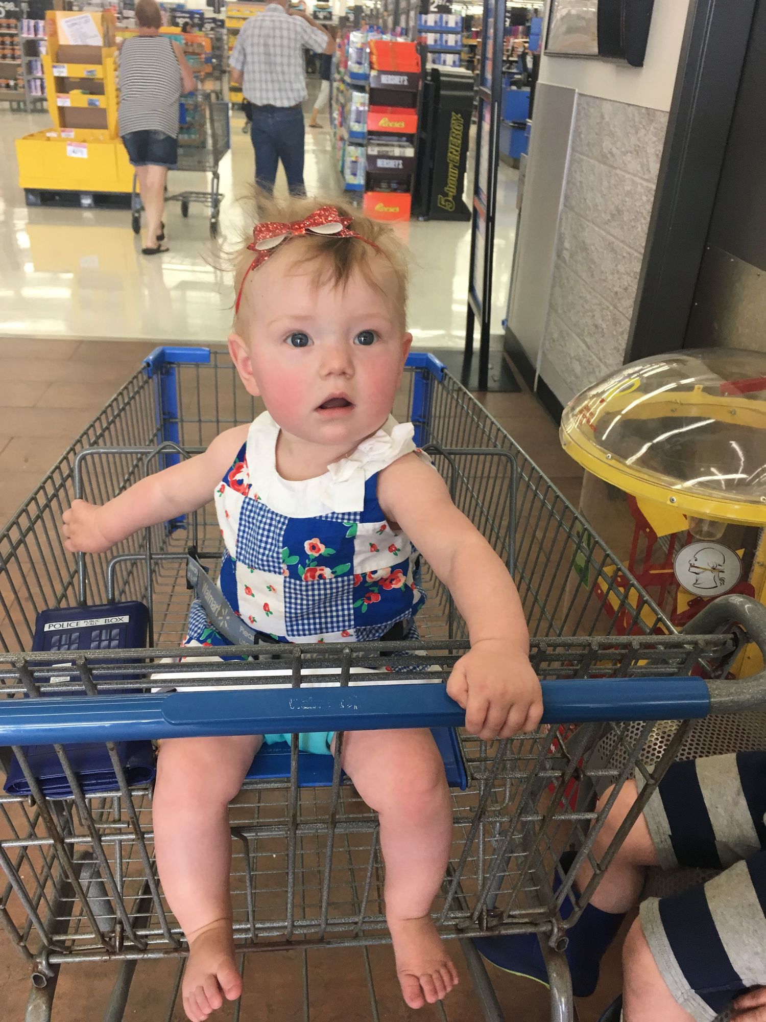 Adorable baby girl sitting in a shopping cart seat at a grocery store wearing a blue romper.