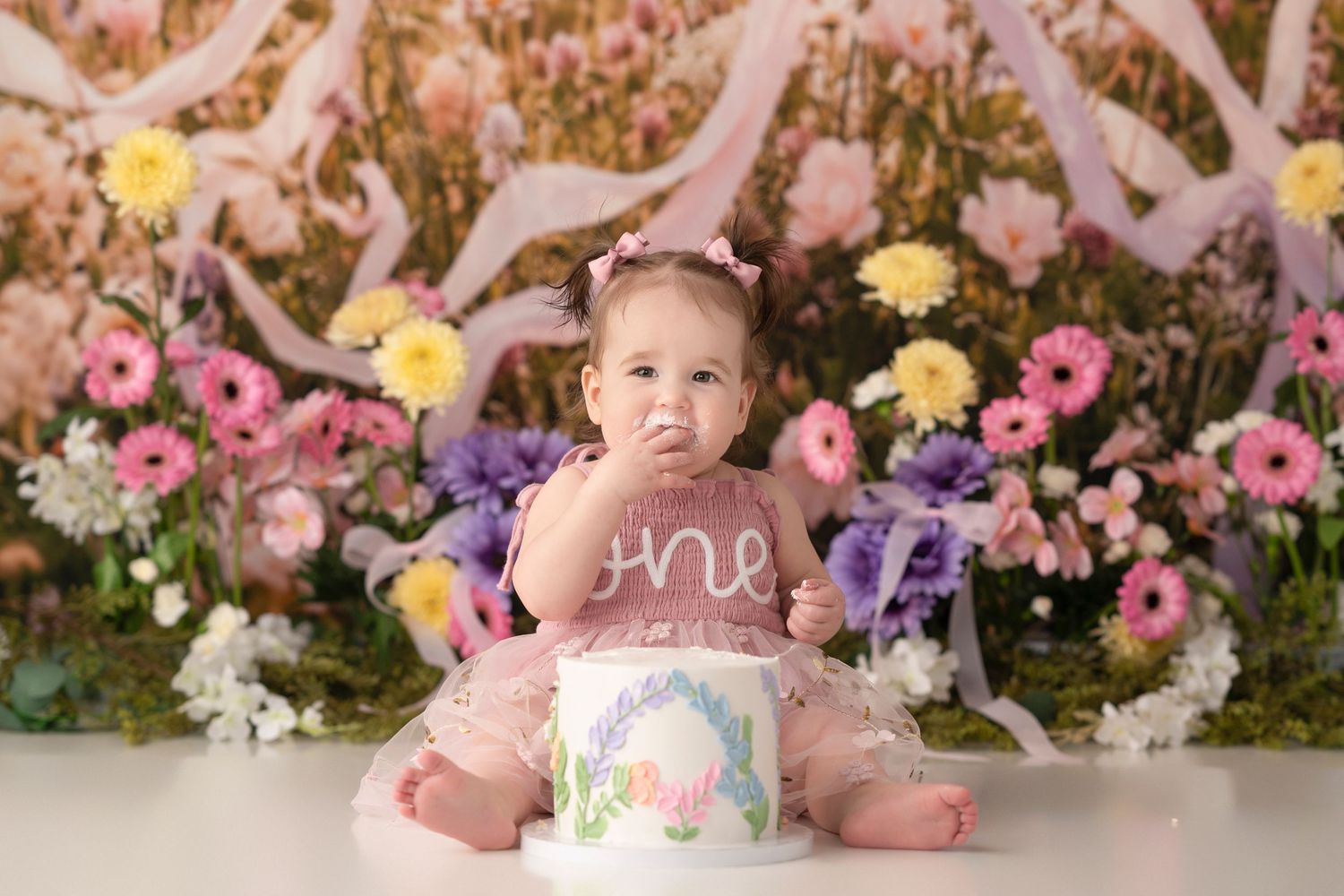 Baby girl in pink tutu sits before a floral backdrop with a white “one” birthday smash cake.