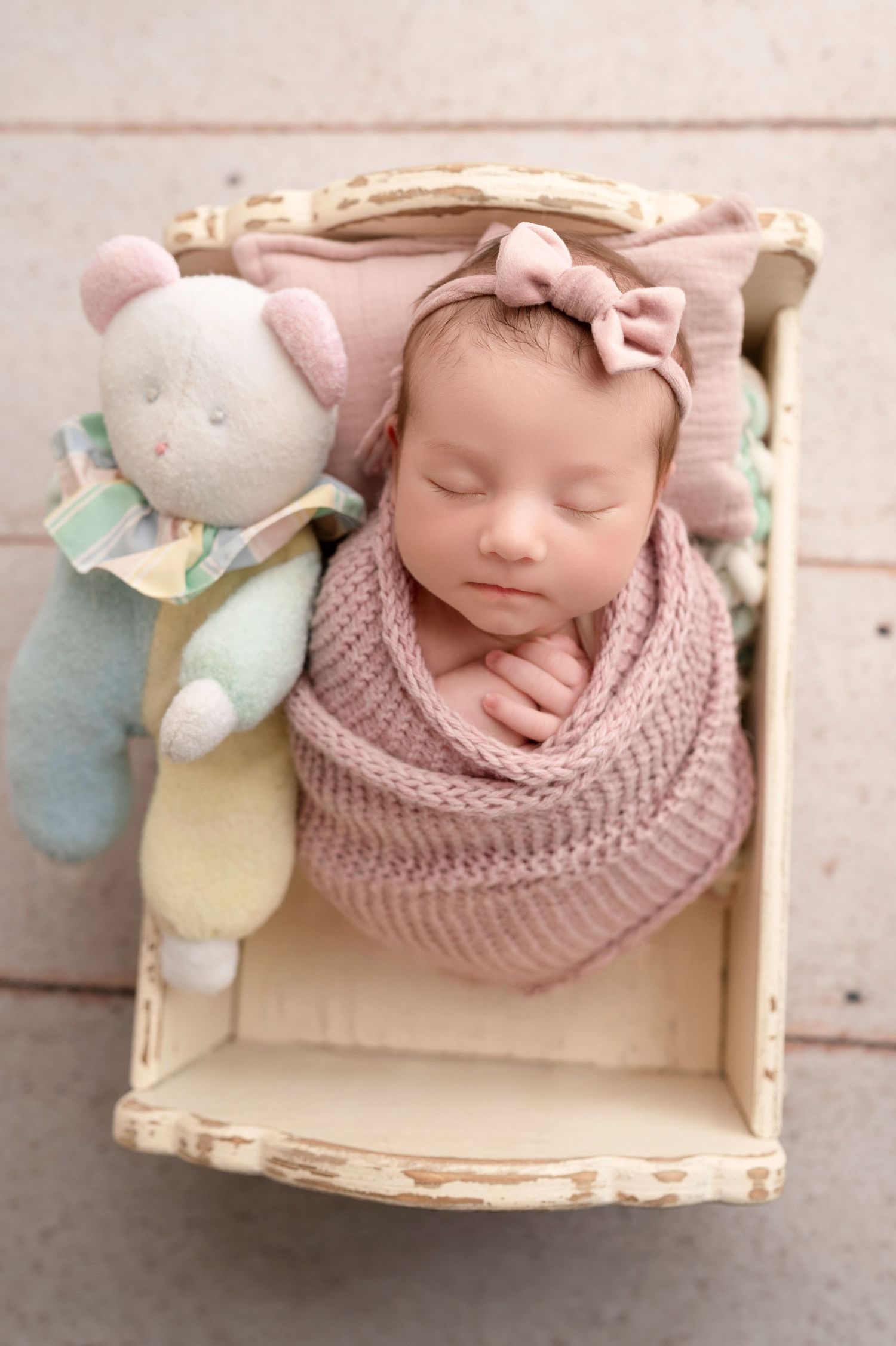 Sleeping newborn baby girl wrapped in pink knit swaddle with bow headband, nestled in vintage wooden crate with stuffed bear.