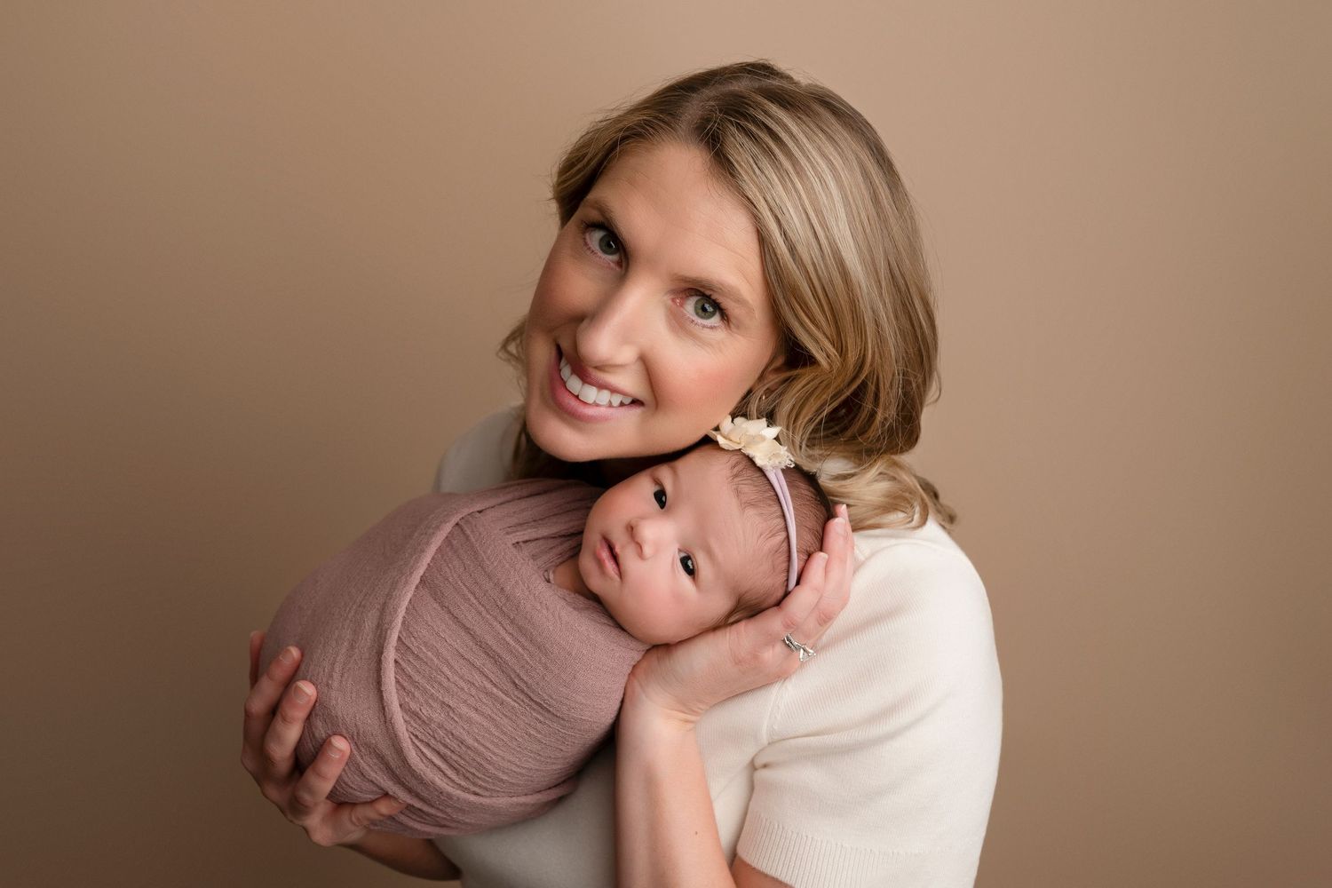 Smiling mother holding swaddled newborn baby in pink wrap with floral headband against beige background.