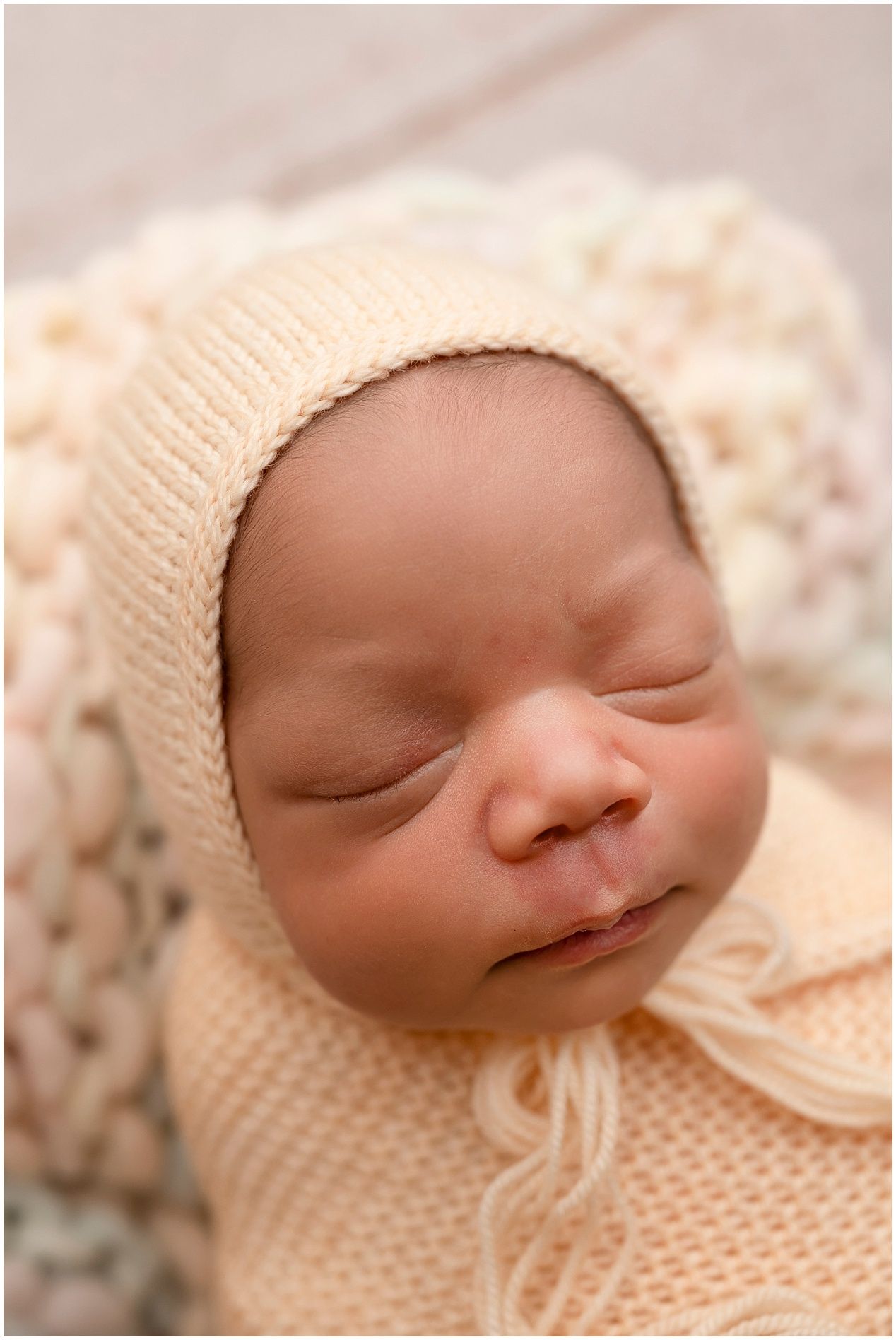 Sleeping newborn baby wrapped in a soft peach knit blanket and matching bonnet hat in a cozy studio portrait.