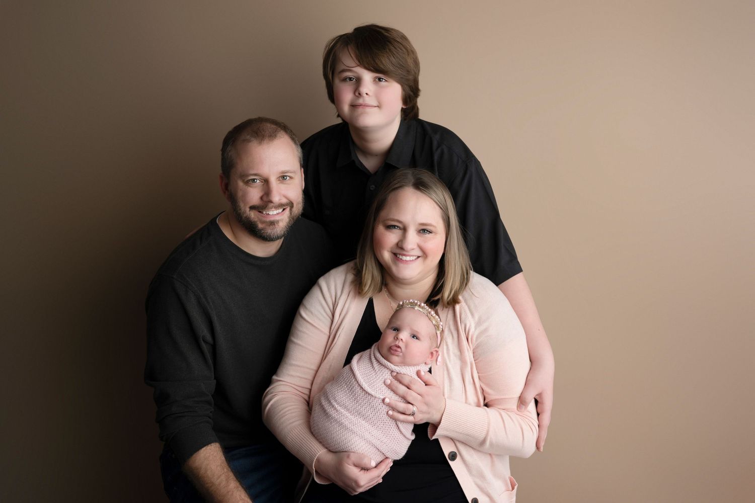 Family portrait with parents, teen son, and newborn baby posed against a warm beige studio background.