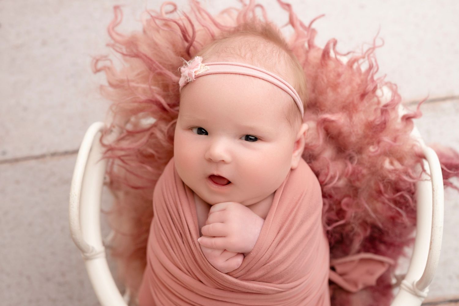 Newborn baby wrapped in pink swaddle with floral headband, nestled in white basket with fluffy pink fur.
