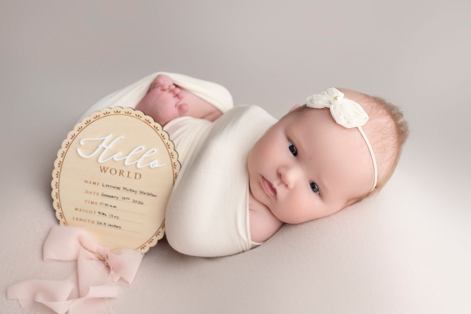 Newborn baby girl swaddled in white wrap with bow headband beside a Hello World birth announcement sign.