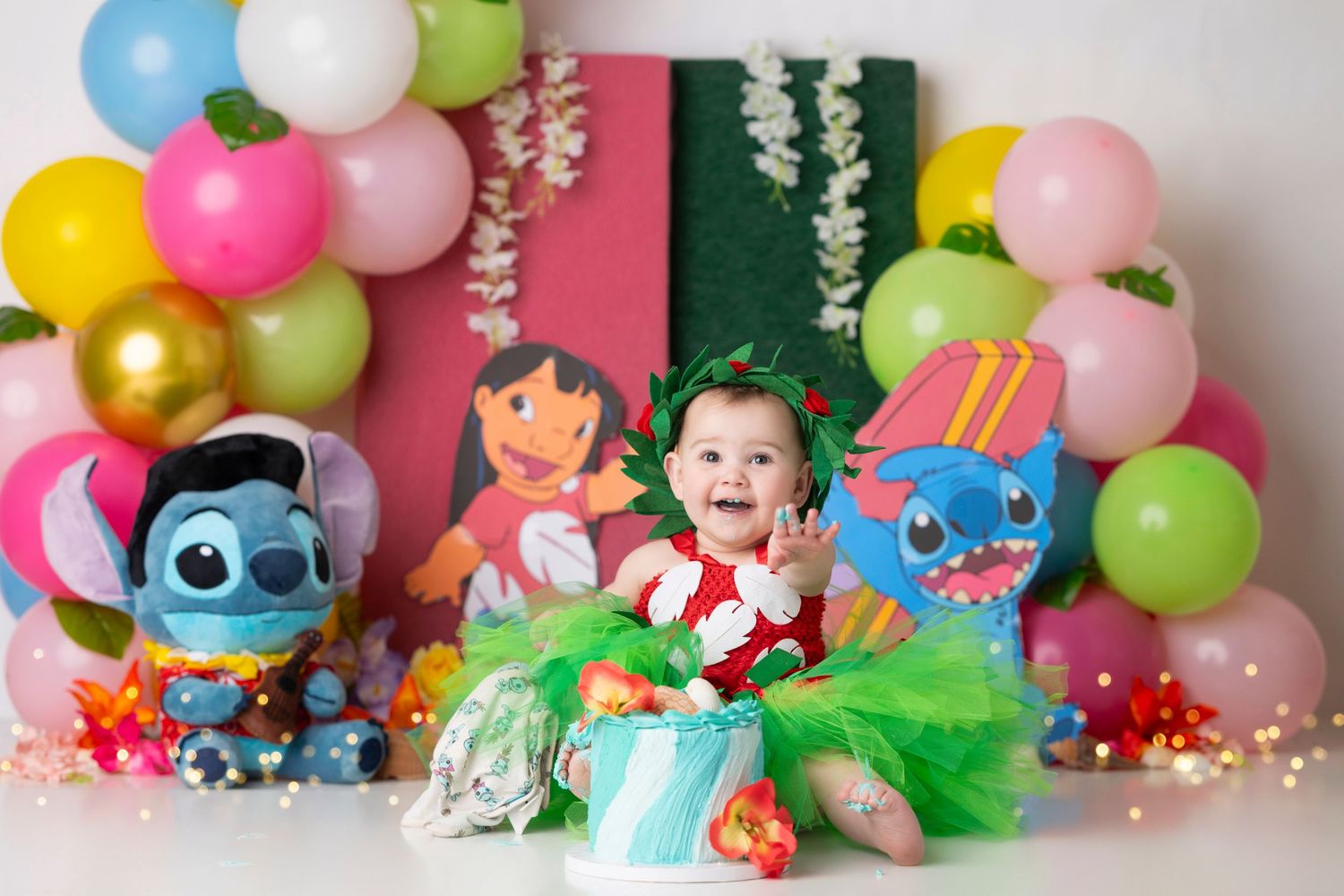 Baby in green tutu sits among Lilo and Stitch plush toys and colorful balloon arch at themed first birthday party.