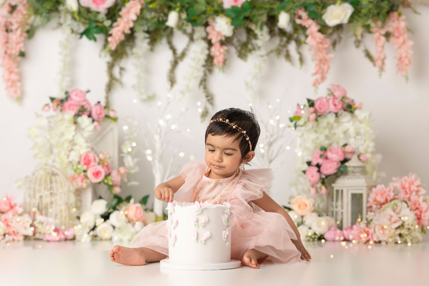 Baby girl in pink tutu touches white cake during floral first birthday cake smash photo session.