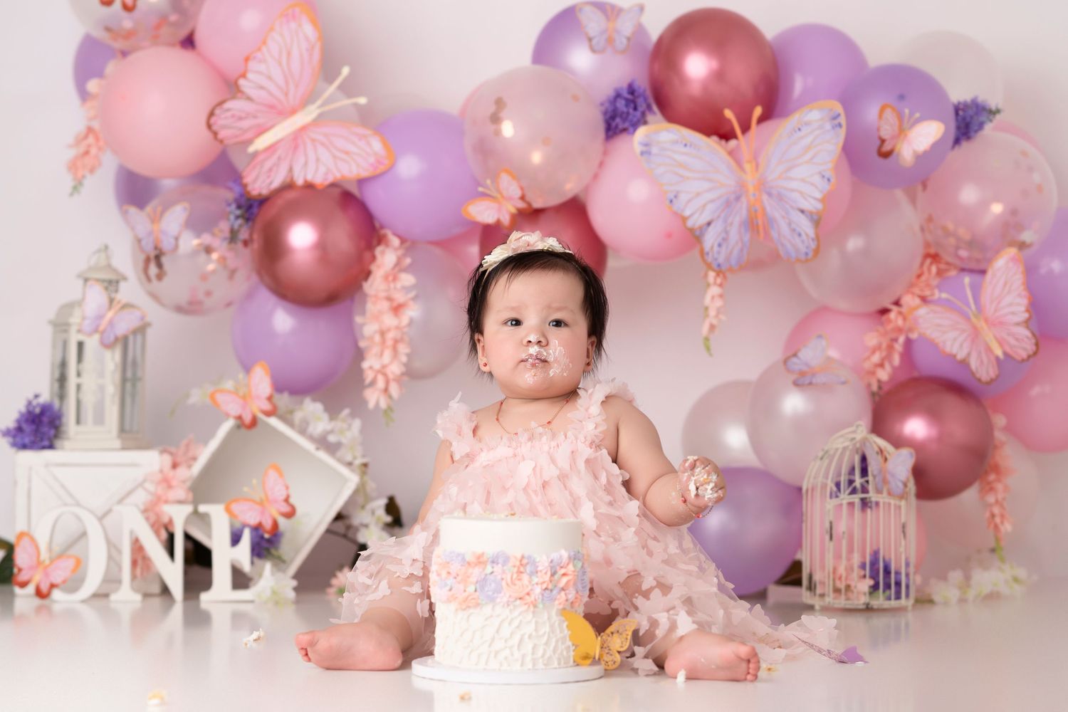 Baby girl in pink dress sits before a first birthday cake, surrounded by butterfly decorations and pink balloons.
