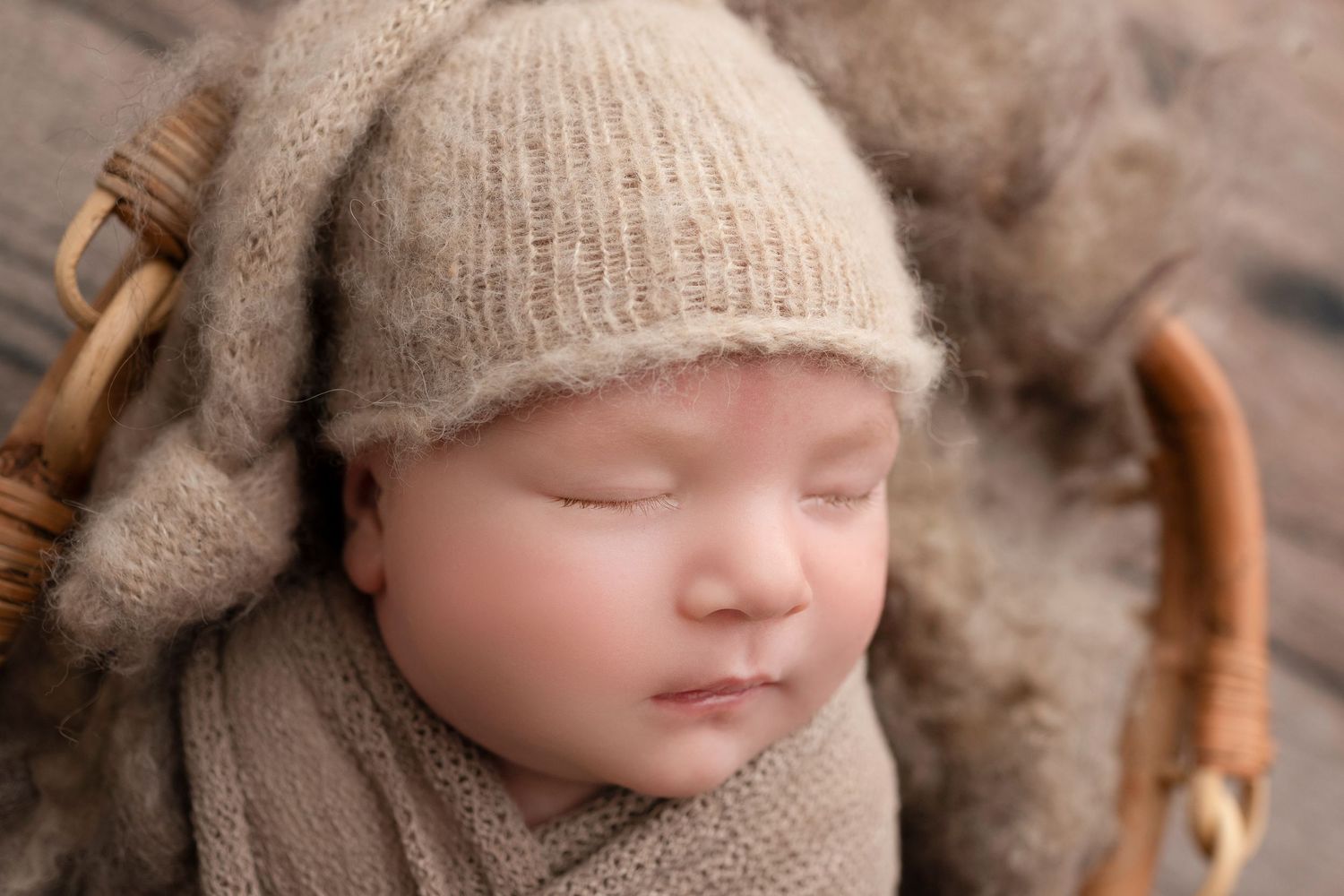 Sleeping newborn baby wrapped in a soft beige knit blanket wearing a matching cozy knit hat in a wicker basket.