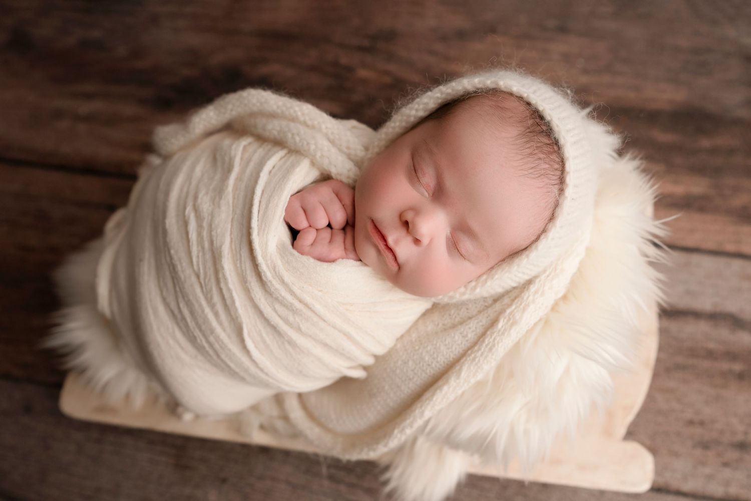 Peaceful newborn wrapped in cream colored blanket sleeping on wooden surface.