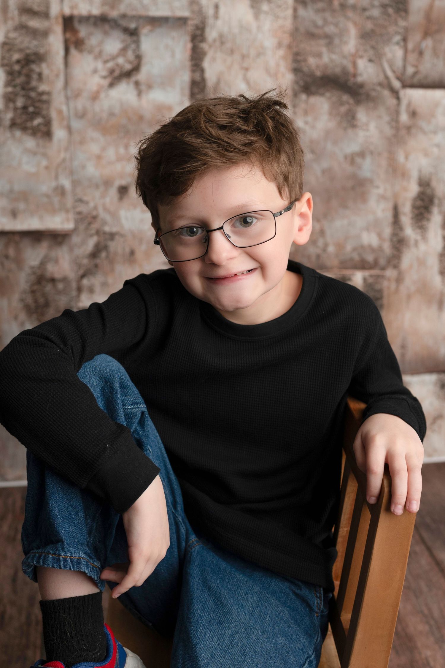 Young person in black sweater and glasses poses casually in wooden chair against textured brown backdrop.