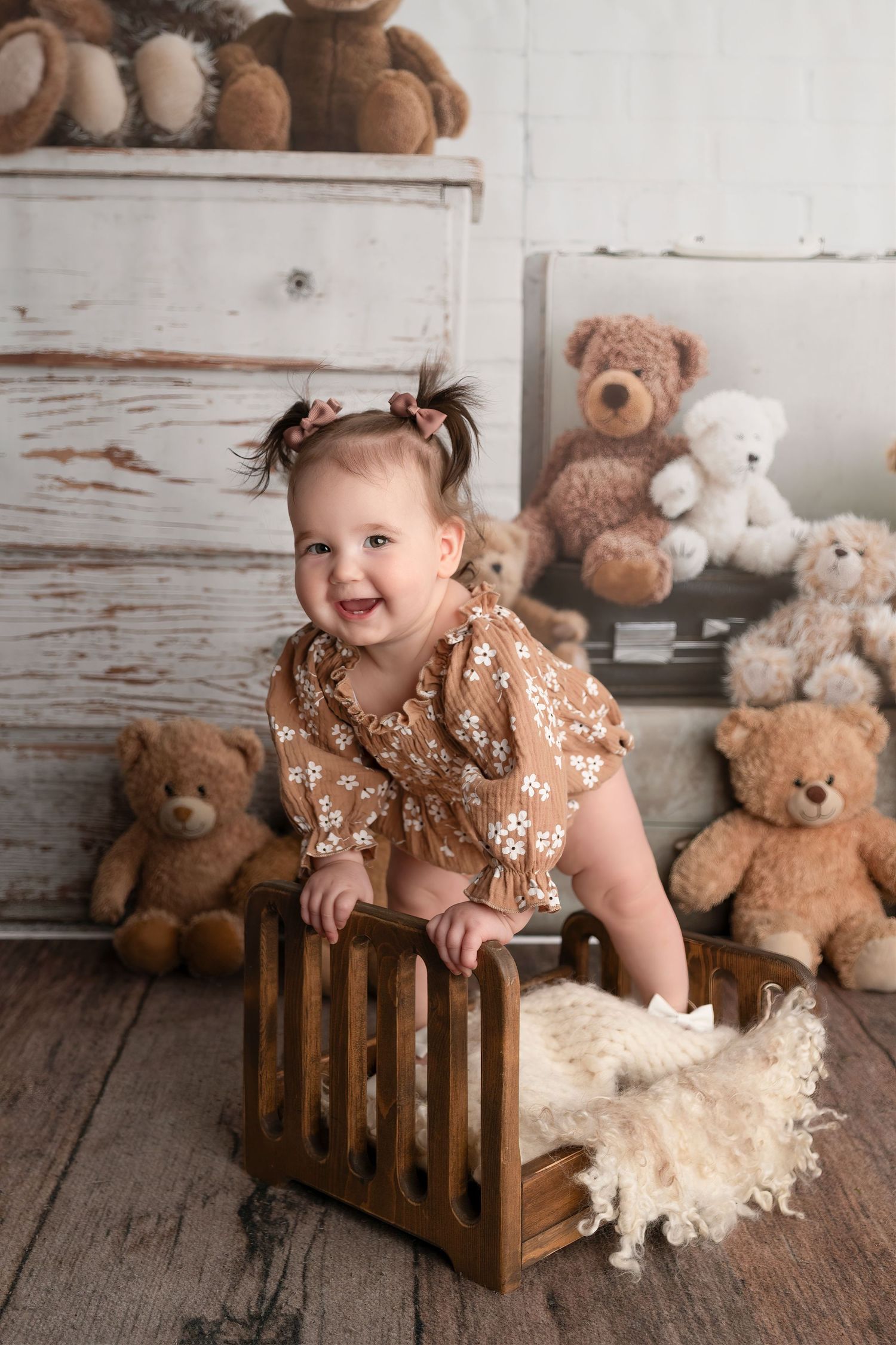 Smiling baby girl in floral romper sitting in wooden crate surrounded by teddy bears in rustic photo studio setting.