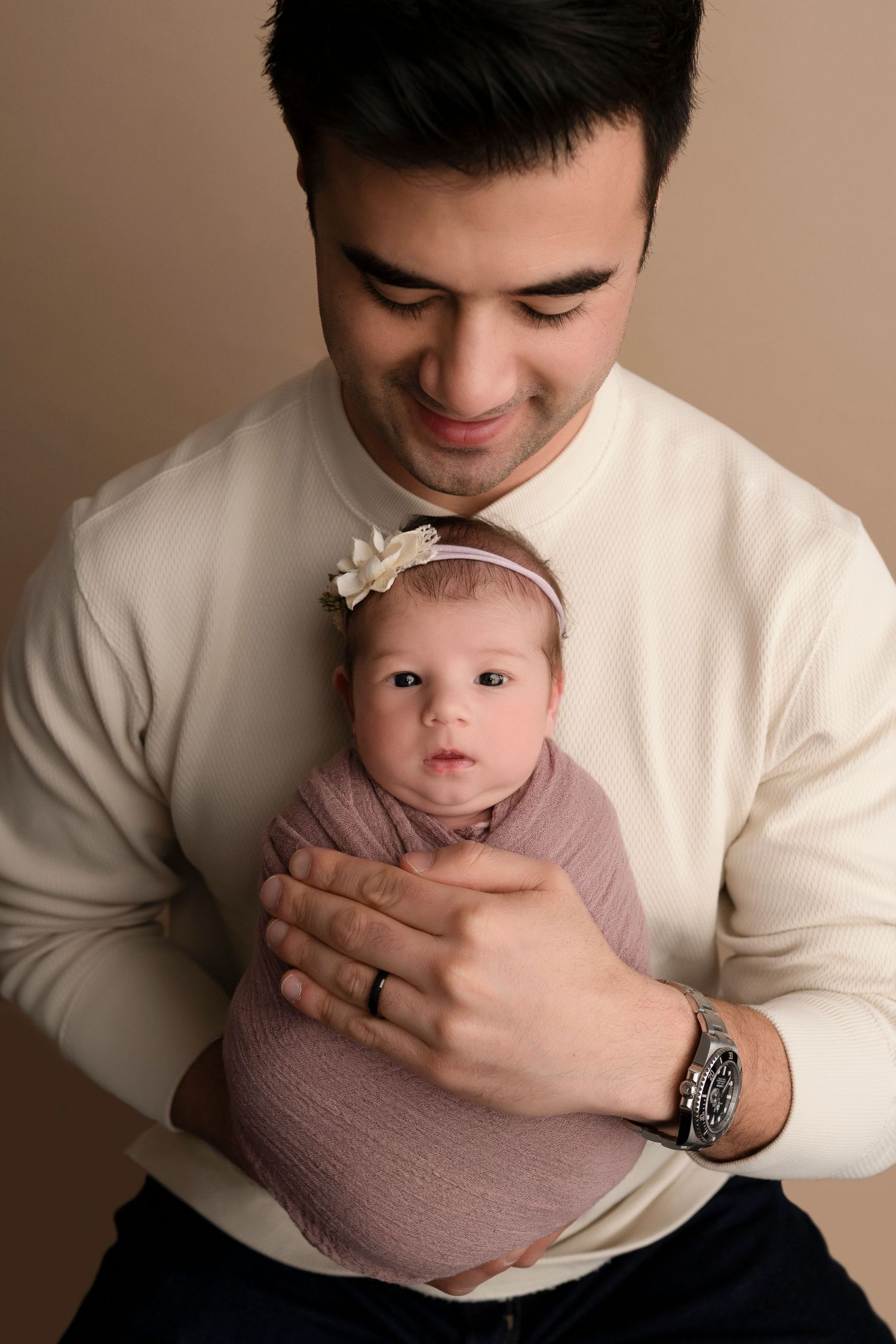 Father in white sweater tenderly holds swaddled newborn baby girl with floral headband against beige background.