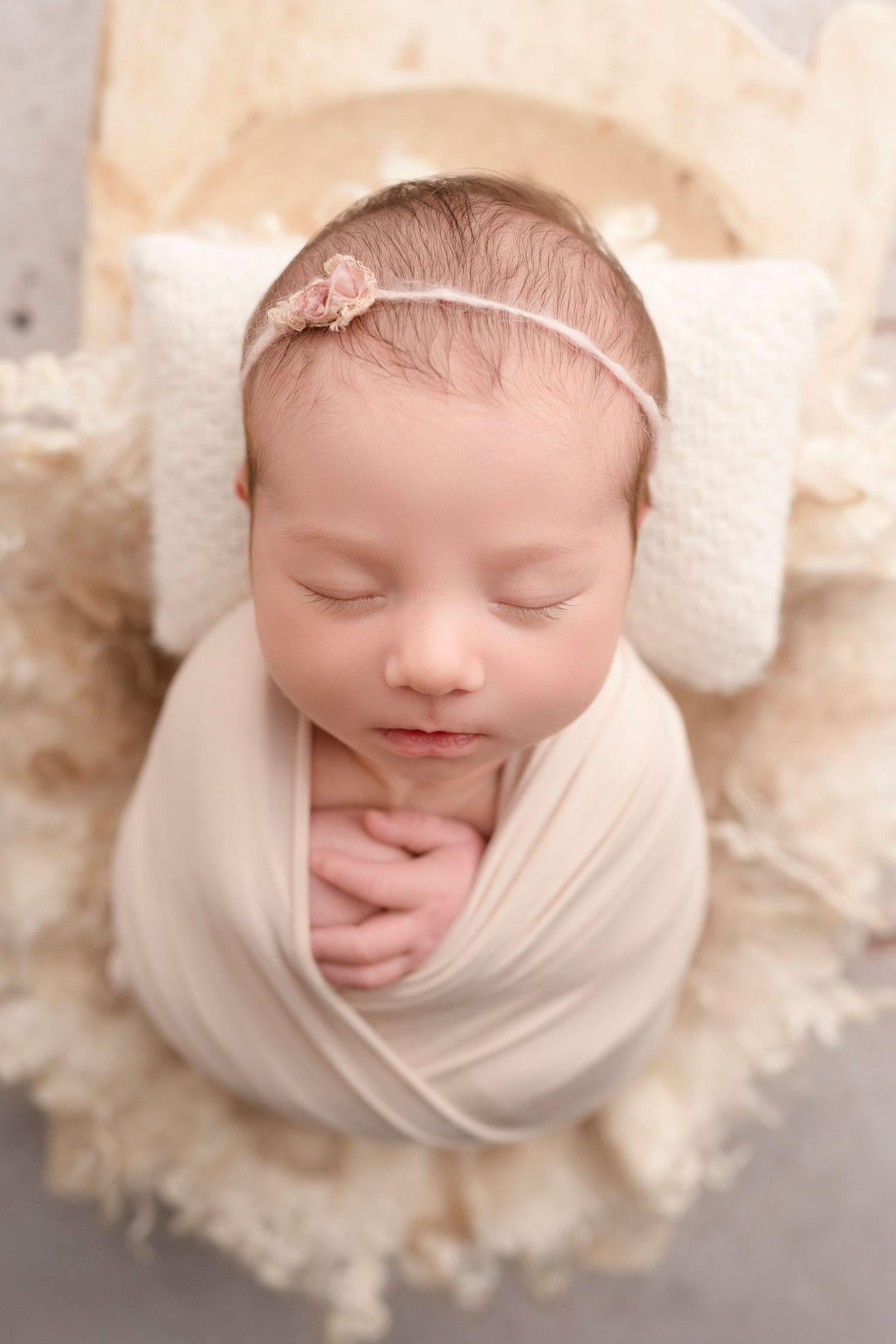 Sleeping newborn baby girl wrapped in cream swaddle with pink floral headband resting on white pillow in wooden bed.