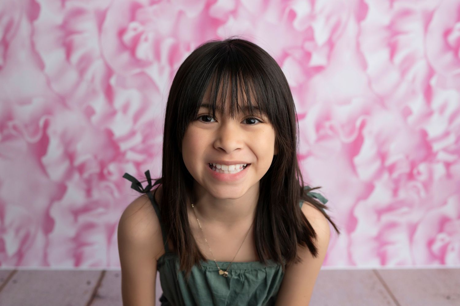 Smiling young girl with dark hair and bangs poses in front of a pink floral backdrop in a studio portrait.