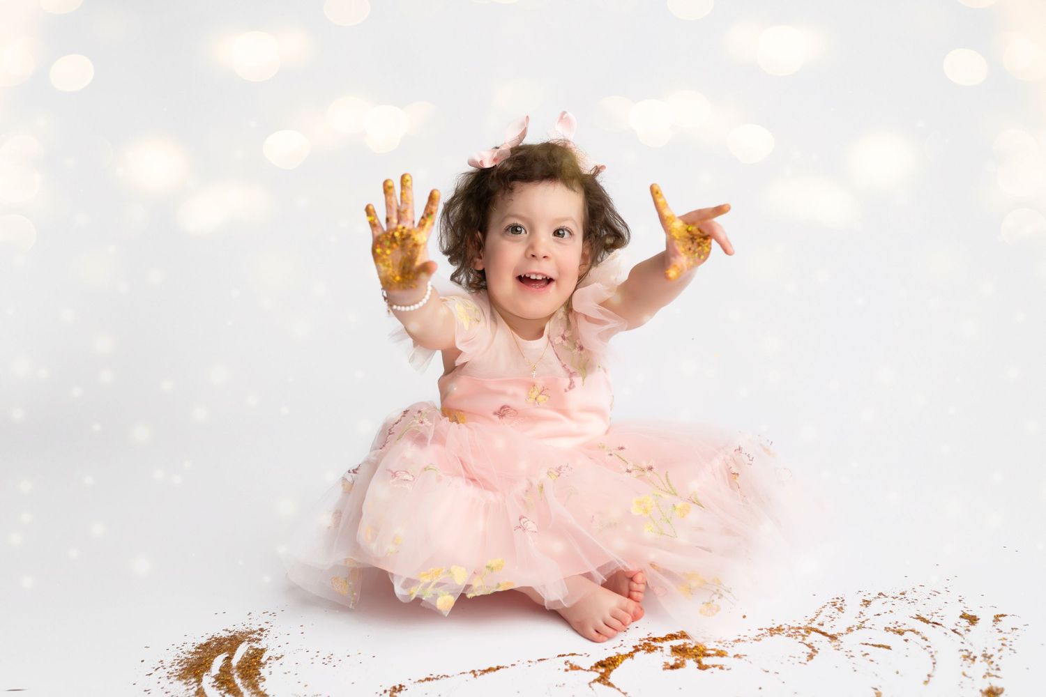 A baby wearing a pink tutu dress sits and plays with gold glitter against a light gray background.