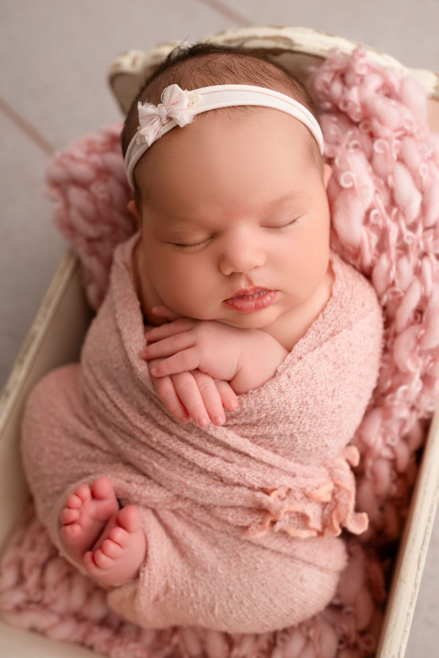 Sleeping newborn wrapped in soft pink blanket with white headband lying in wooden crate with textured pink backdrop.