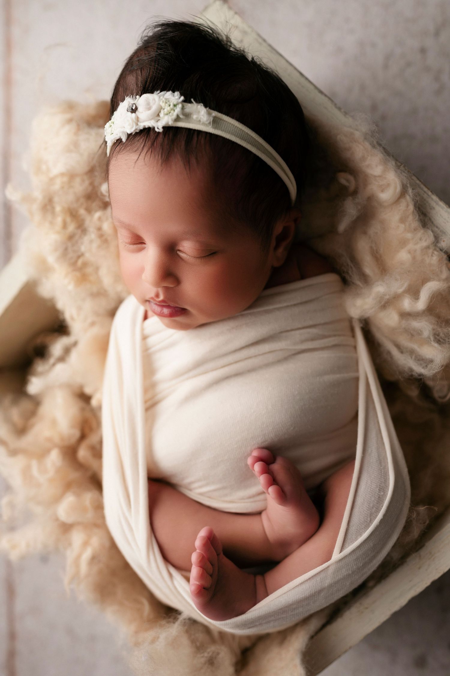 A sleeping newborn baby wrapped in soft white fabric lies peacefully on a cream-colored textured background.