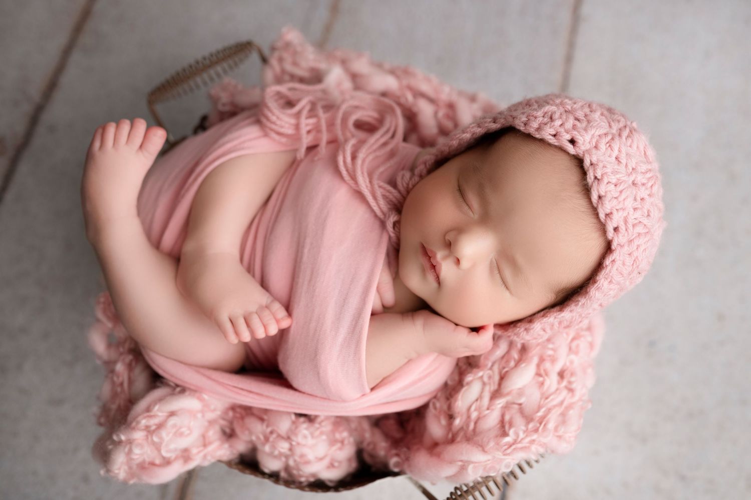 Newborn baby sleeping peacefully in soft pink knit wrap on textured gray background.