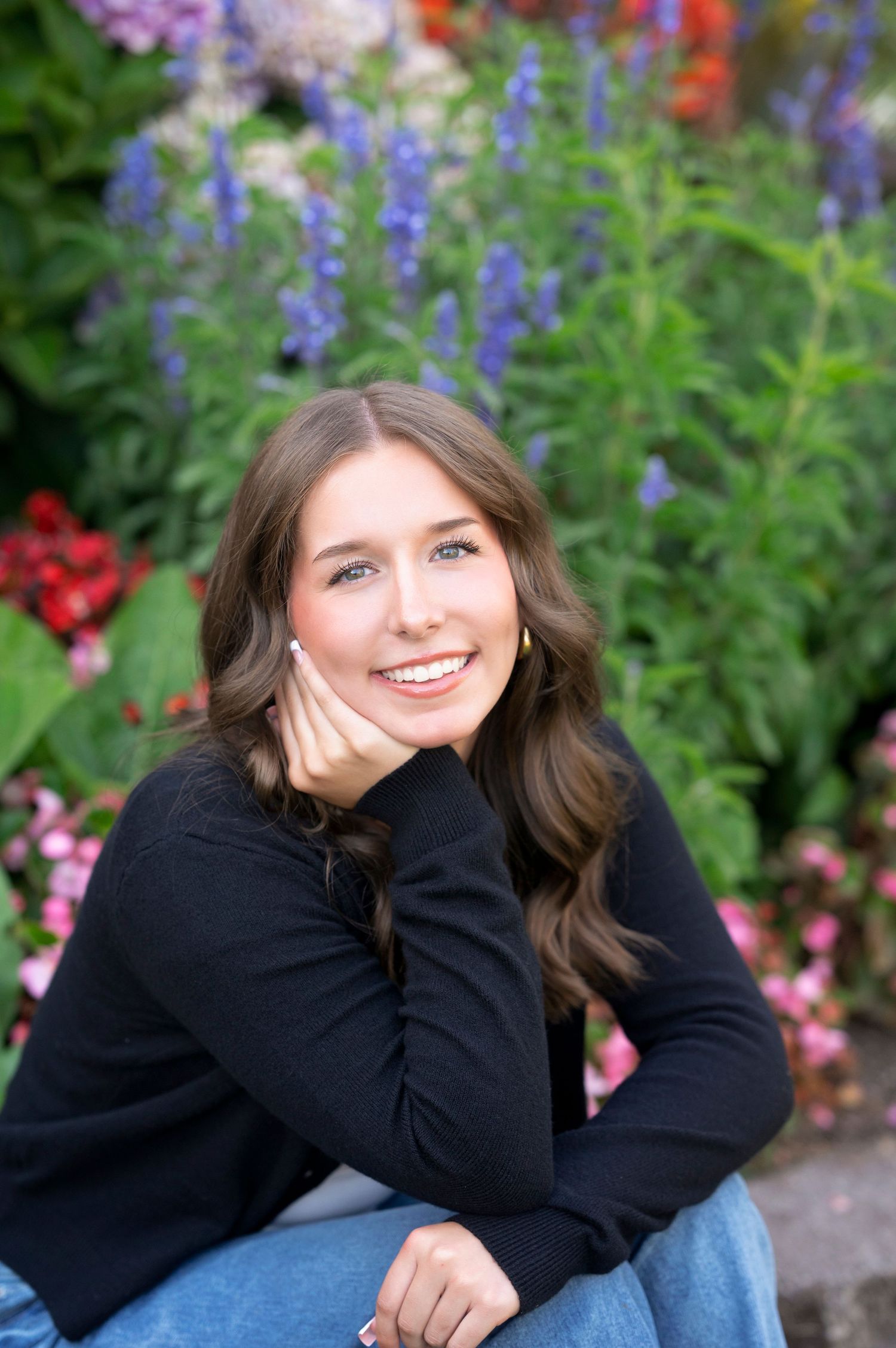 Smiling person in black sweater sits casually in front of colorful garden with purple and red flowers.