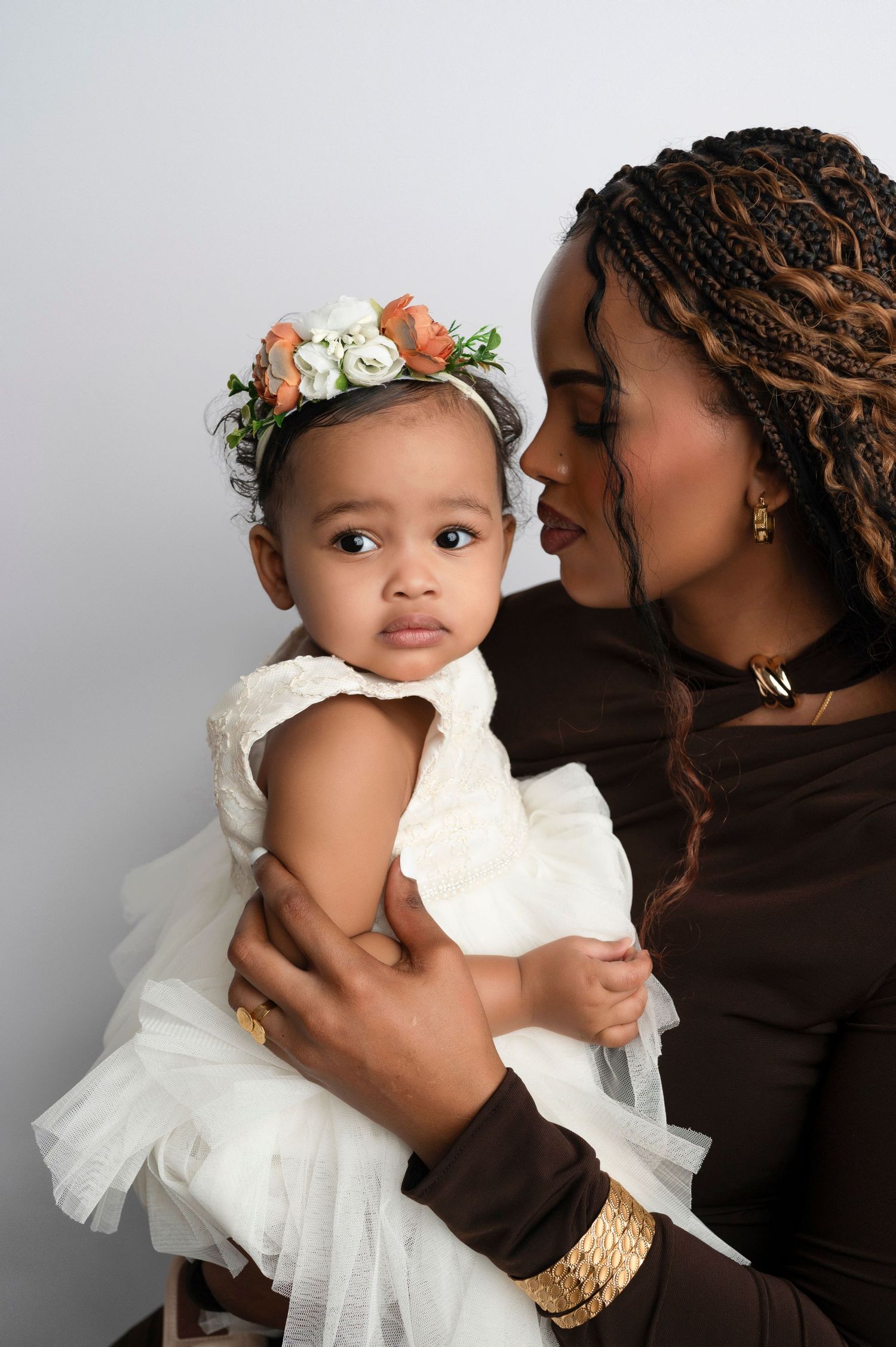 A Black mother in brown dress holds her baby girl wearing white tulle dress and floral crown in a studio portrait.