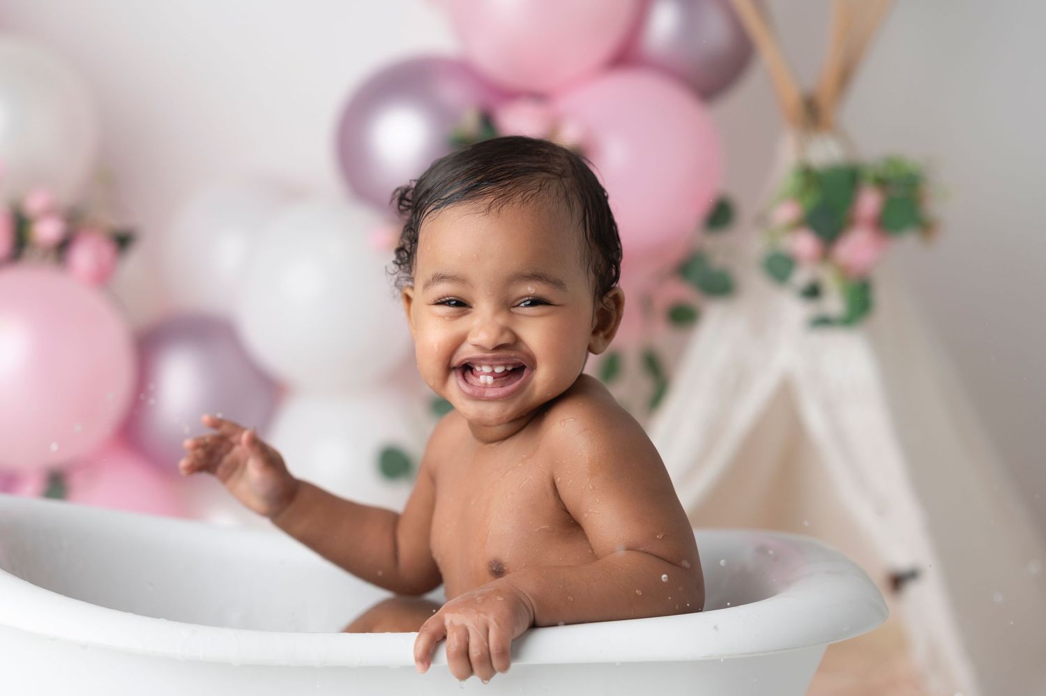 A joyful baby smiling in a white bathtub surrounded by pink and white balloons in a bright studio setting.