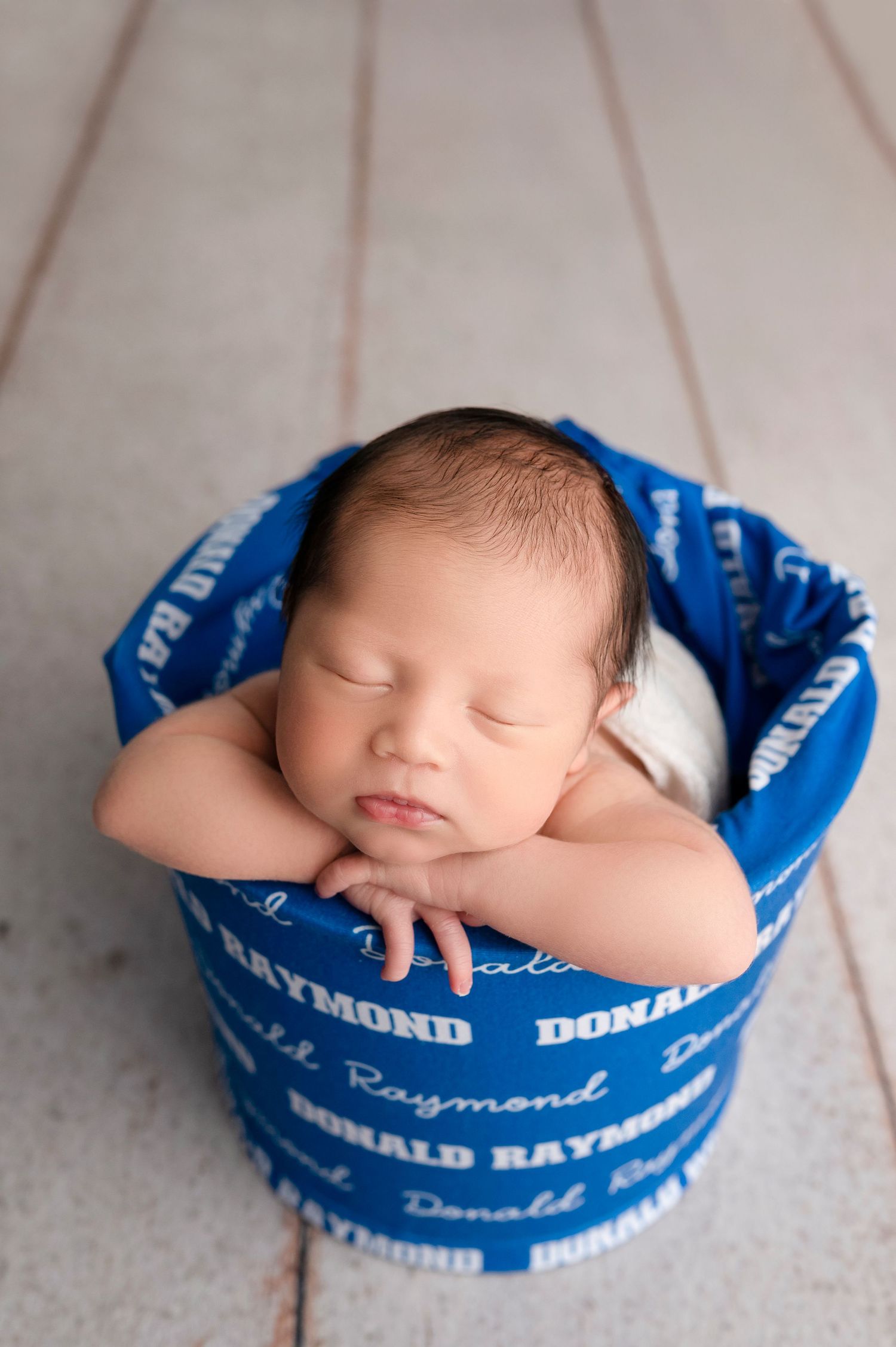Sleeping newborn baby resting in a blue bucket wrap printed with the name Donald Raymond for a professional newborn photo.