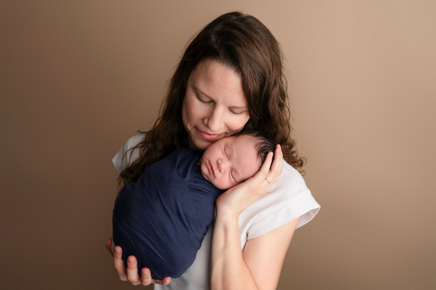 Mother tenderly holds sleeping newborn wrapped in navy blue swaddle against her cheek in a studio portrait.