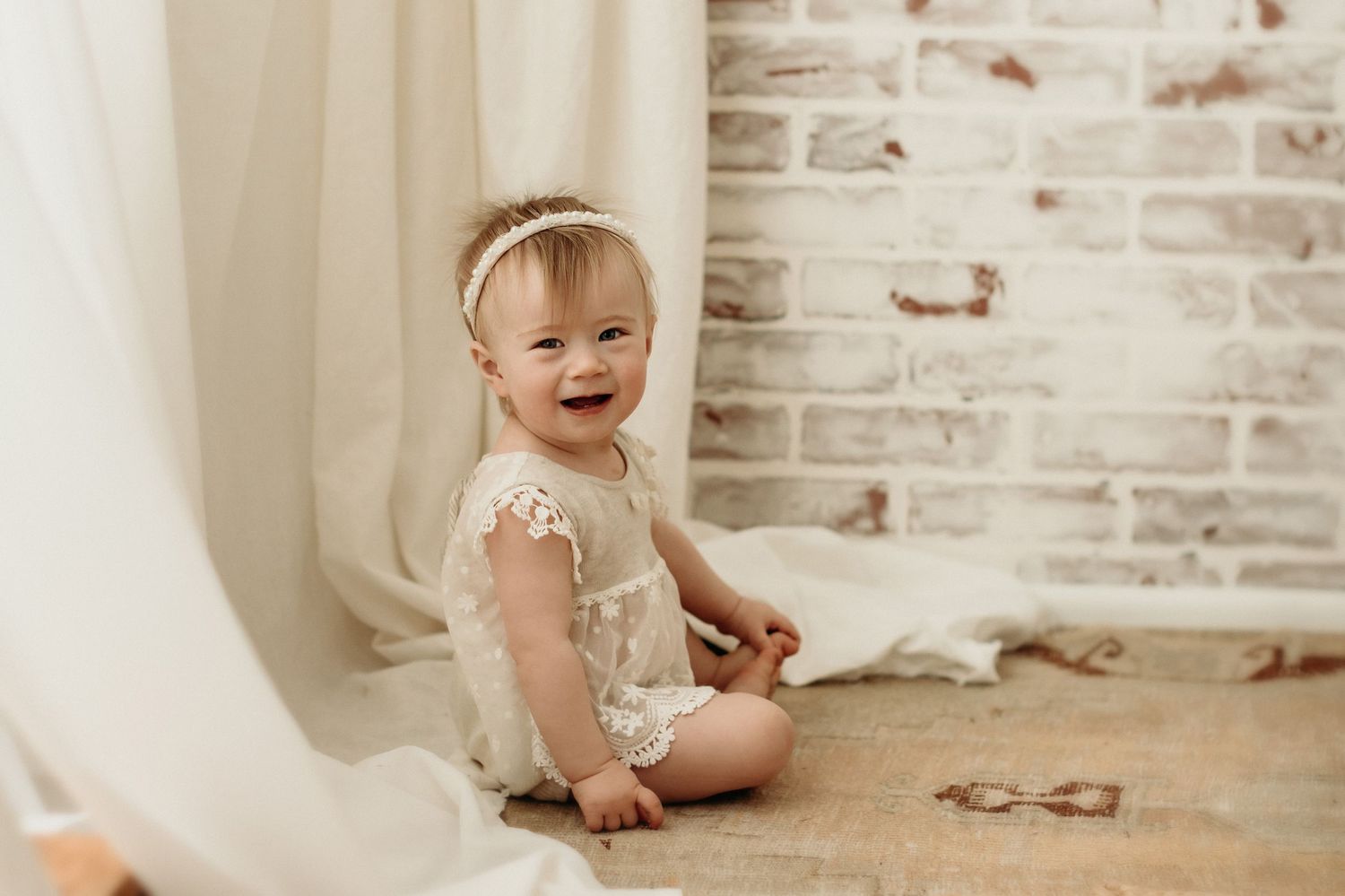 A cheerful baby in a lace dress sits on hardwood flooring against a white brick wall backdrop.