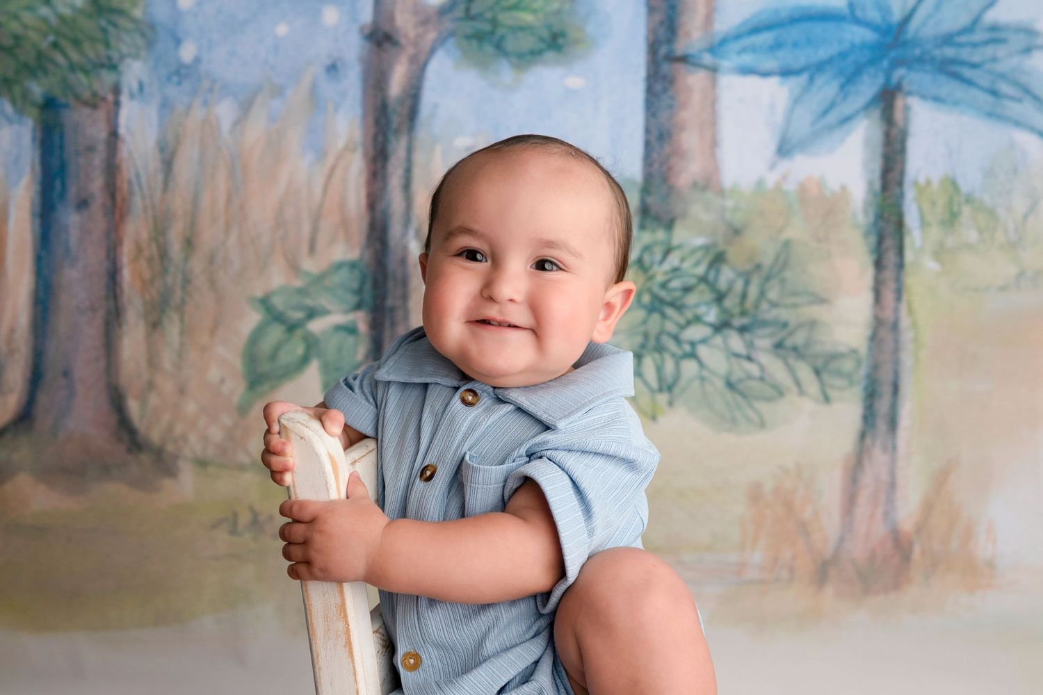 Smiling baby in blue romper sitting on white chair with painted forest backdrop.