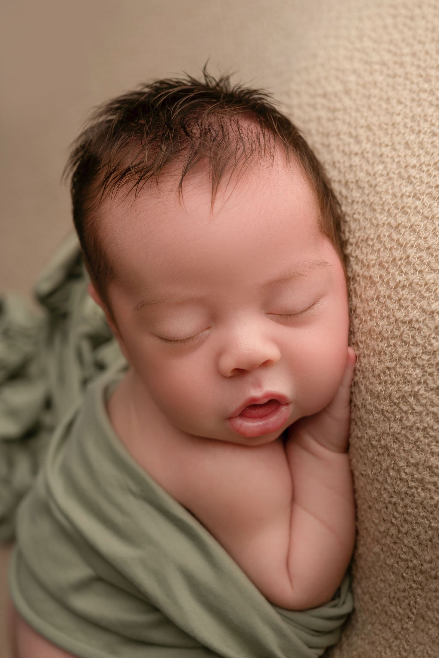 Sleeping newborn baby wrapped in green swaddle cloth resting peacefully against a beige background.