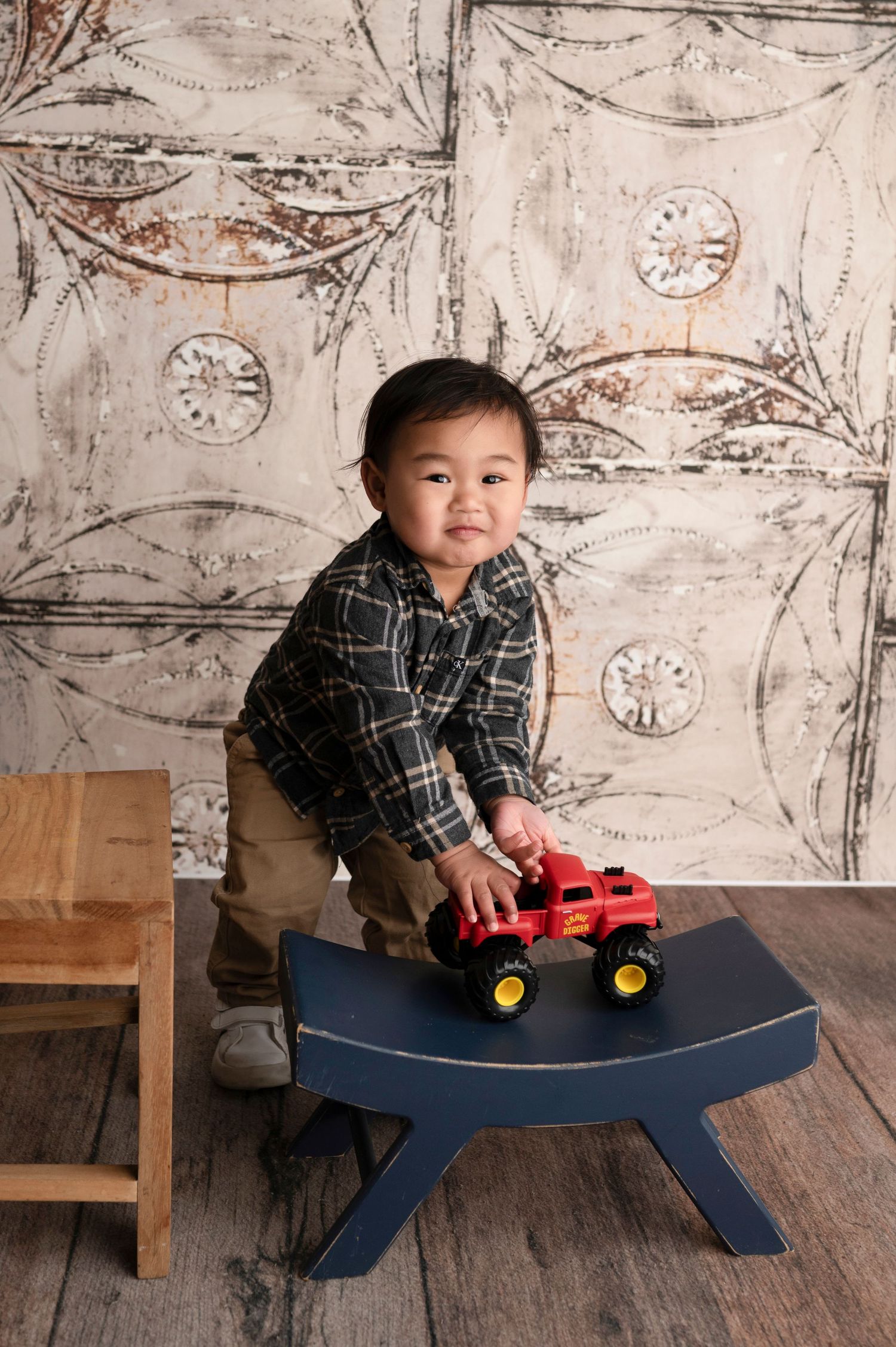Smiling toddler boy in plaid shirt playing with red monster truck toy on blue step stool with vintage backdrop.