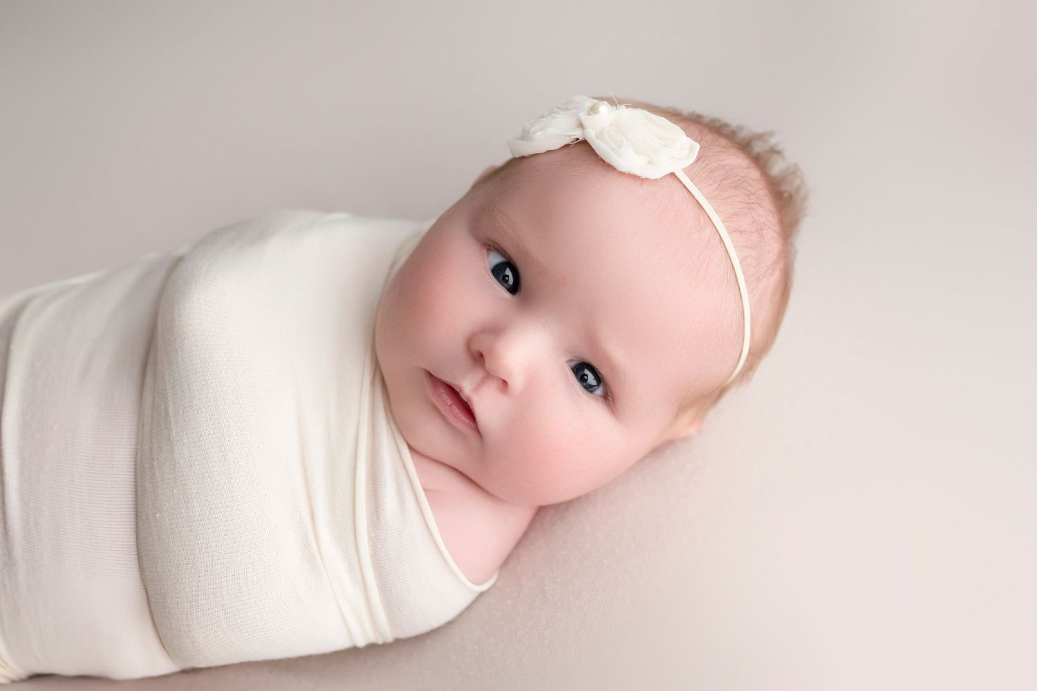 Newborn baby swaddled in white wrap wearing a delicate floral headband, lying peacefully on a soft gray background.
