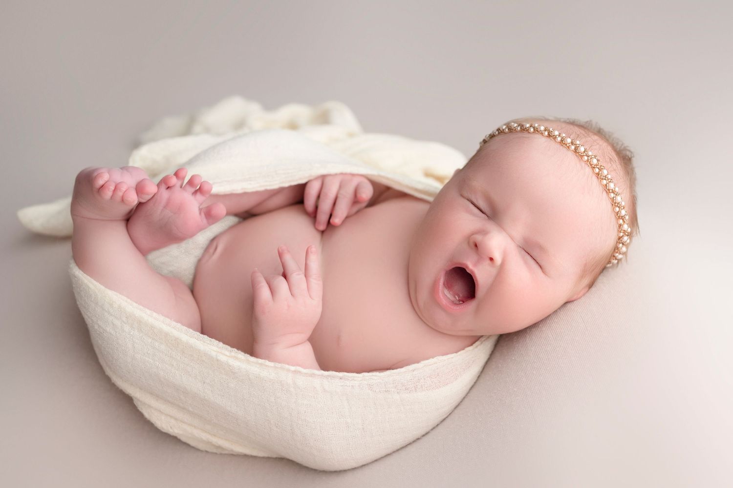 Adorable newborn baby yawning while nestled in a white bowl with a pearl headband during a studio photo session.