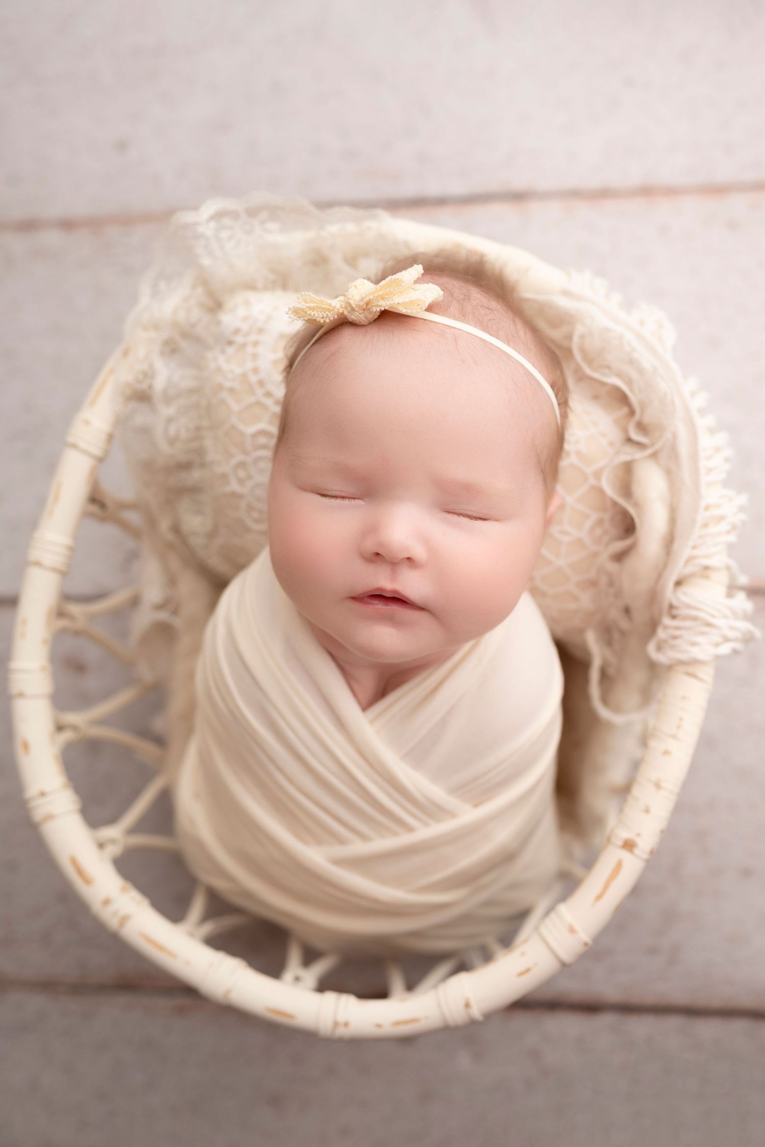 Sleeping newborn baby girl wrapped in cream swaddle with bow headband, nestled in white wicker basket.